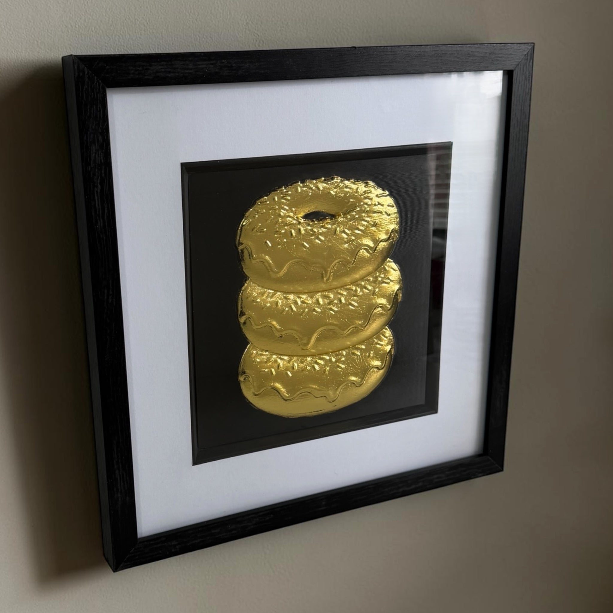 Angled Gold doughnut stack framed artwork showing raised relief detail, black background and glass front.