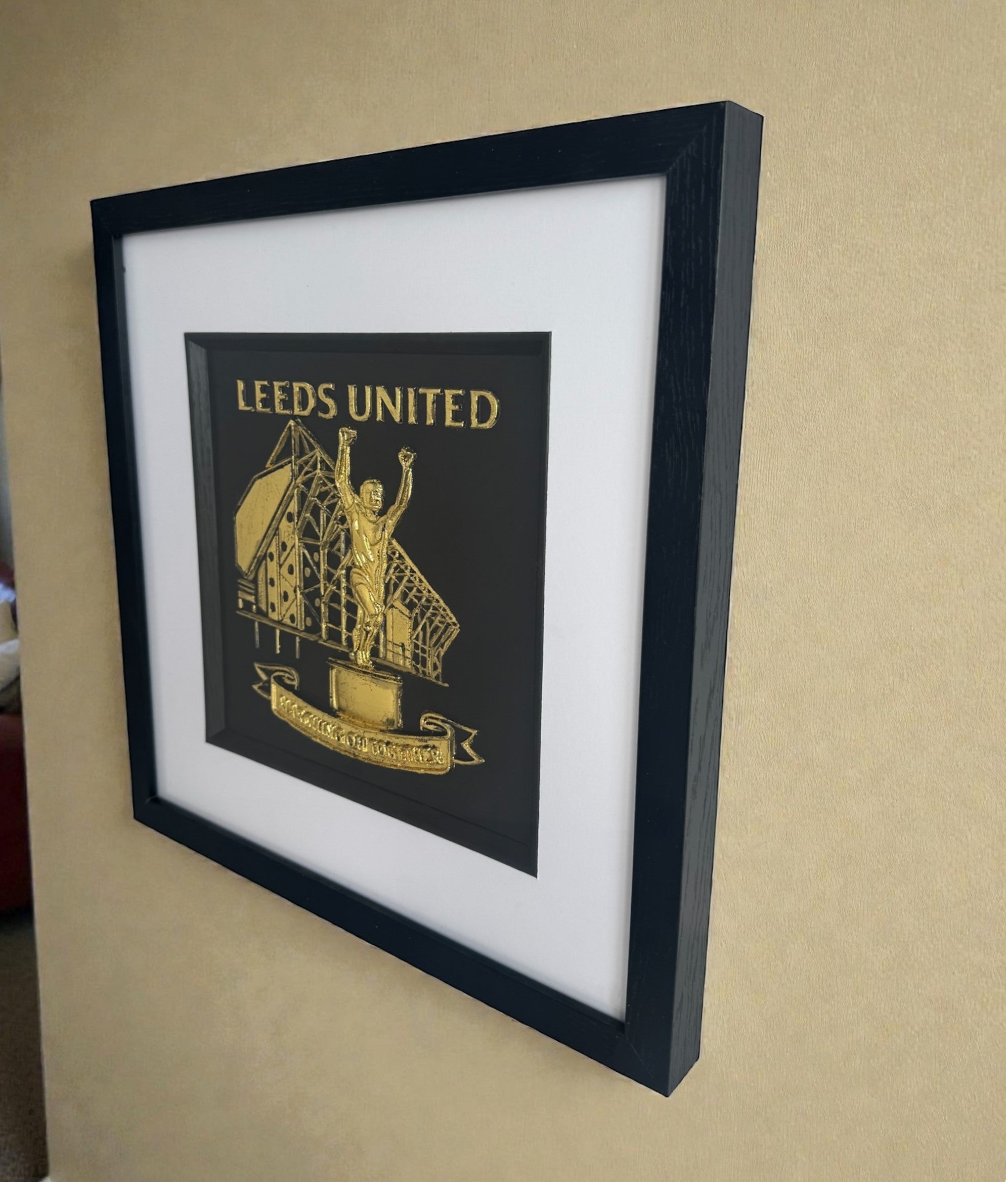 Framed Leeds United 3D relief artwork in Gold Leaf with black box frame, white mount and inner black surround, angled on a neutral wall