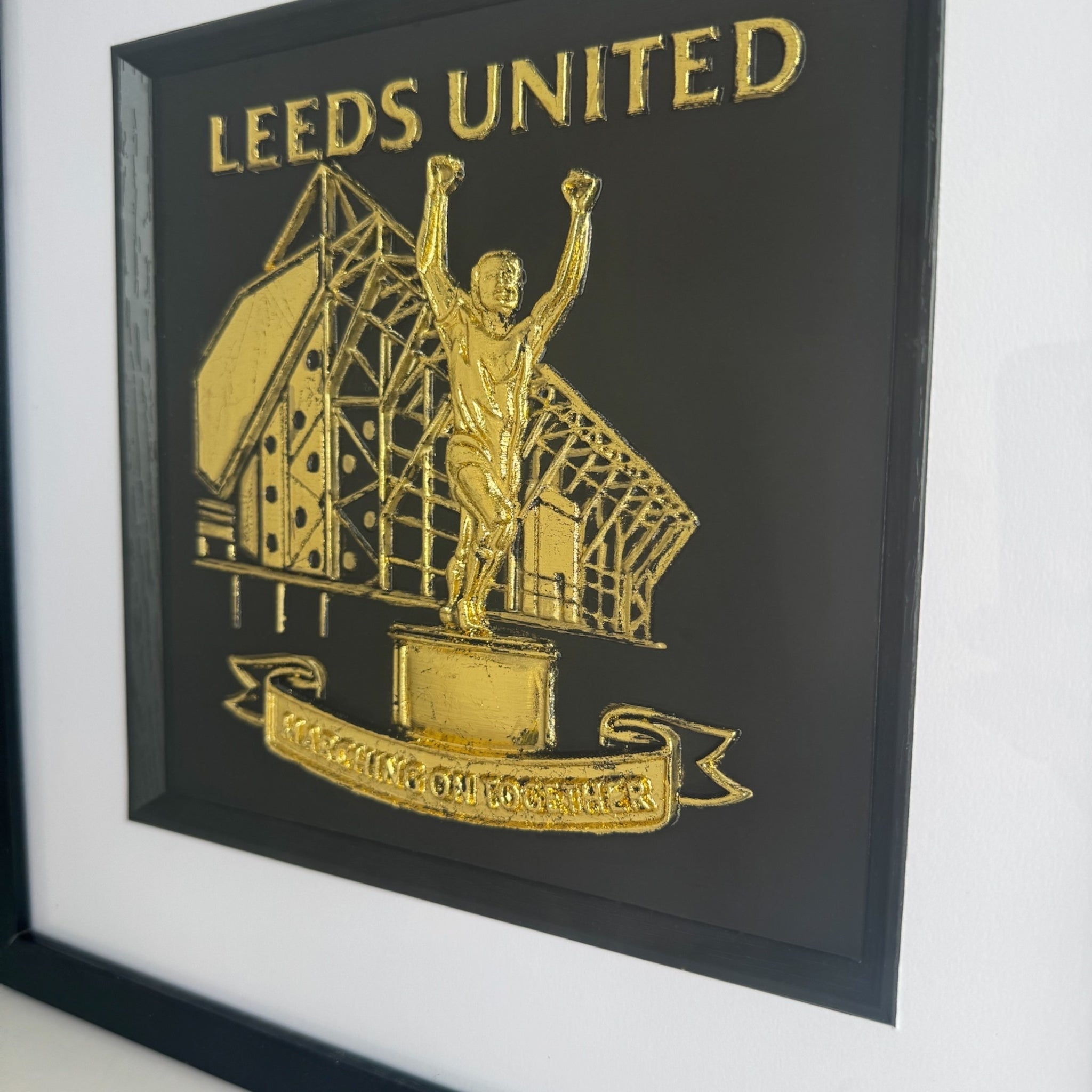 Close-up of Leeds United 3D relief artwork in Gold Leaf showing the Billy Bremner statue and Elland Road detail on a black background with raised “Leeds United” lettering and the “Marching On Together” banner inside a black inner surround.