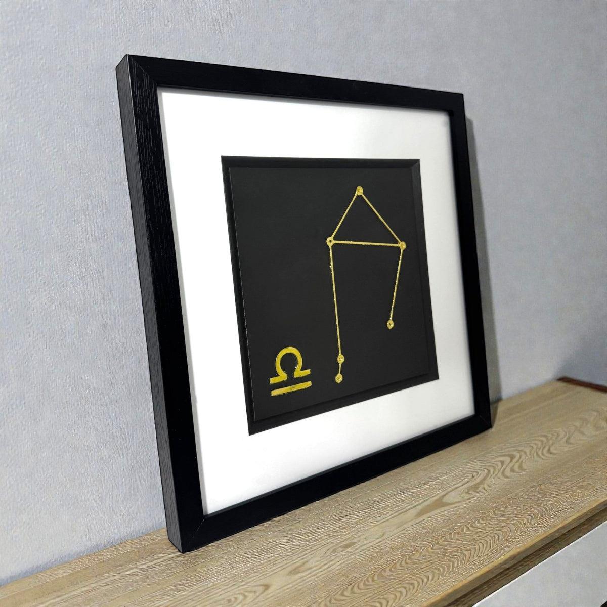 Gold Libra constellation wall art shown at an angle on a wooden surface, framed with white mount and raised zodiac star design