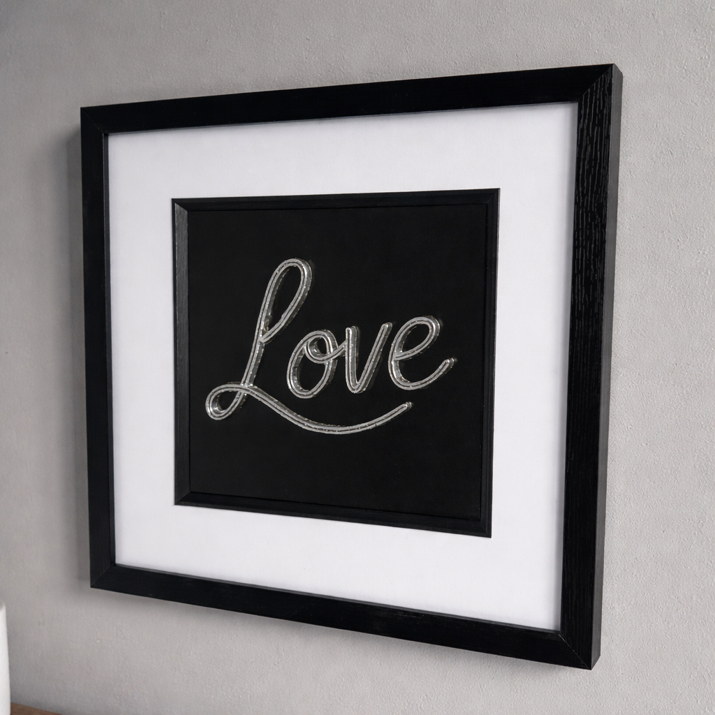 Angled wall display of framed 3D Love sign in silver finish, showing depth and shadow on a grey wall.