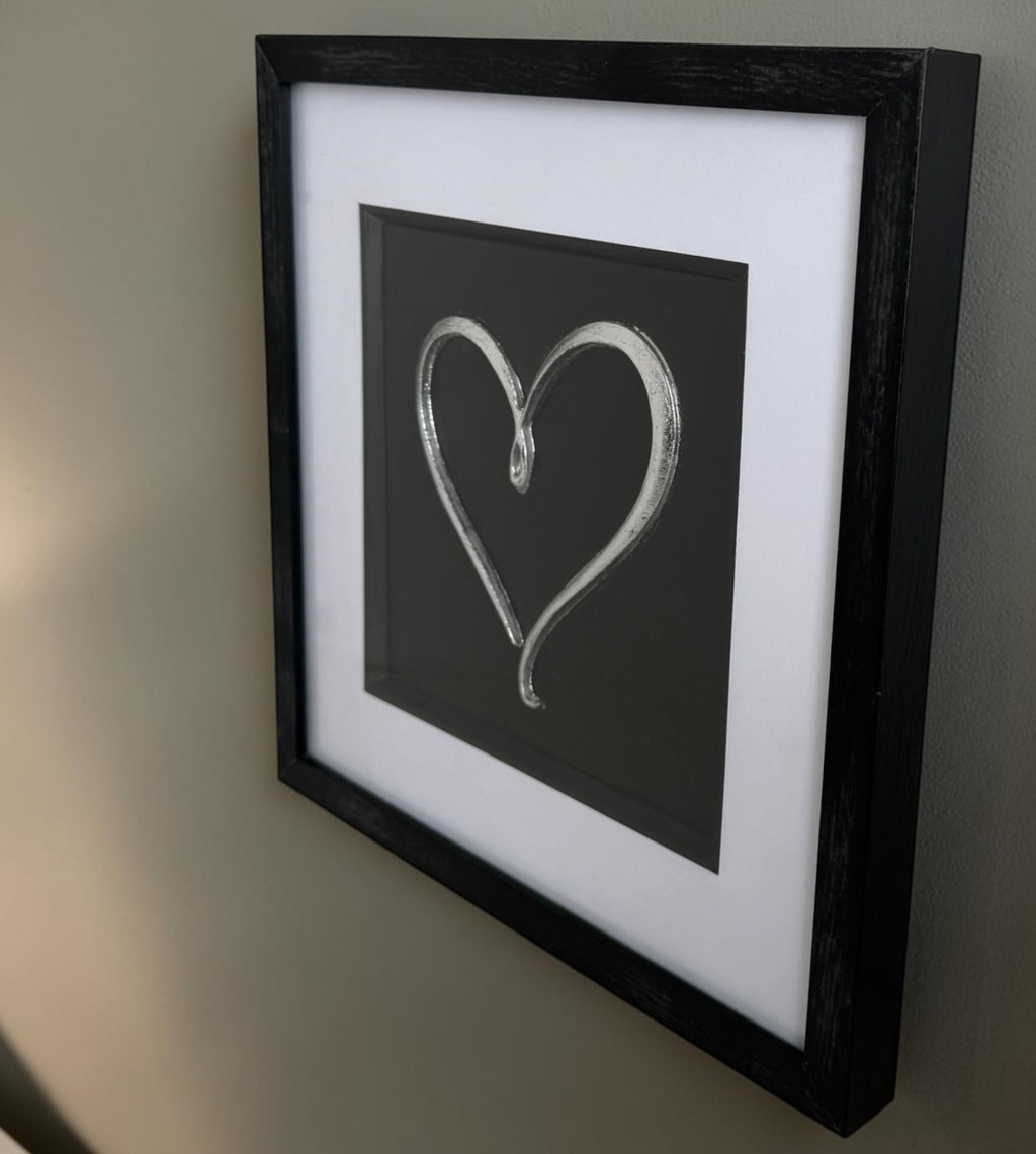 Angled Silver love heart framed artwork showing raised relief detail, glass reflection and black box frame.