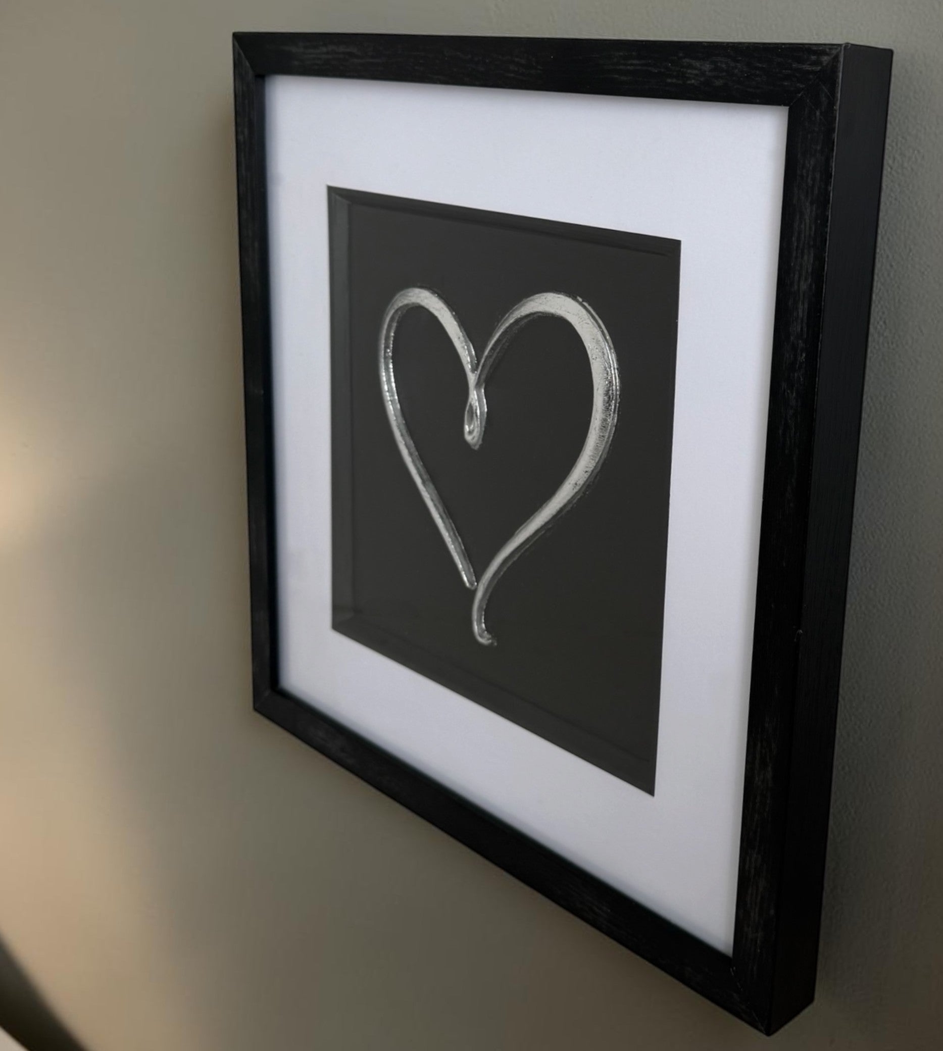 Angled Silver love heart framed artwork showing raised relief detail, glass reflection and black box frame.