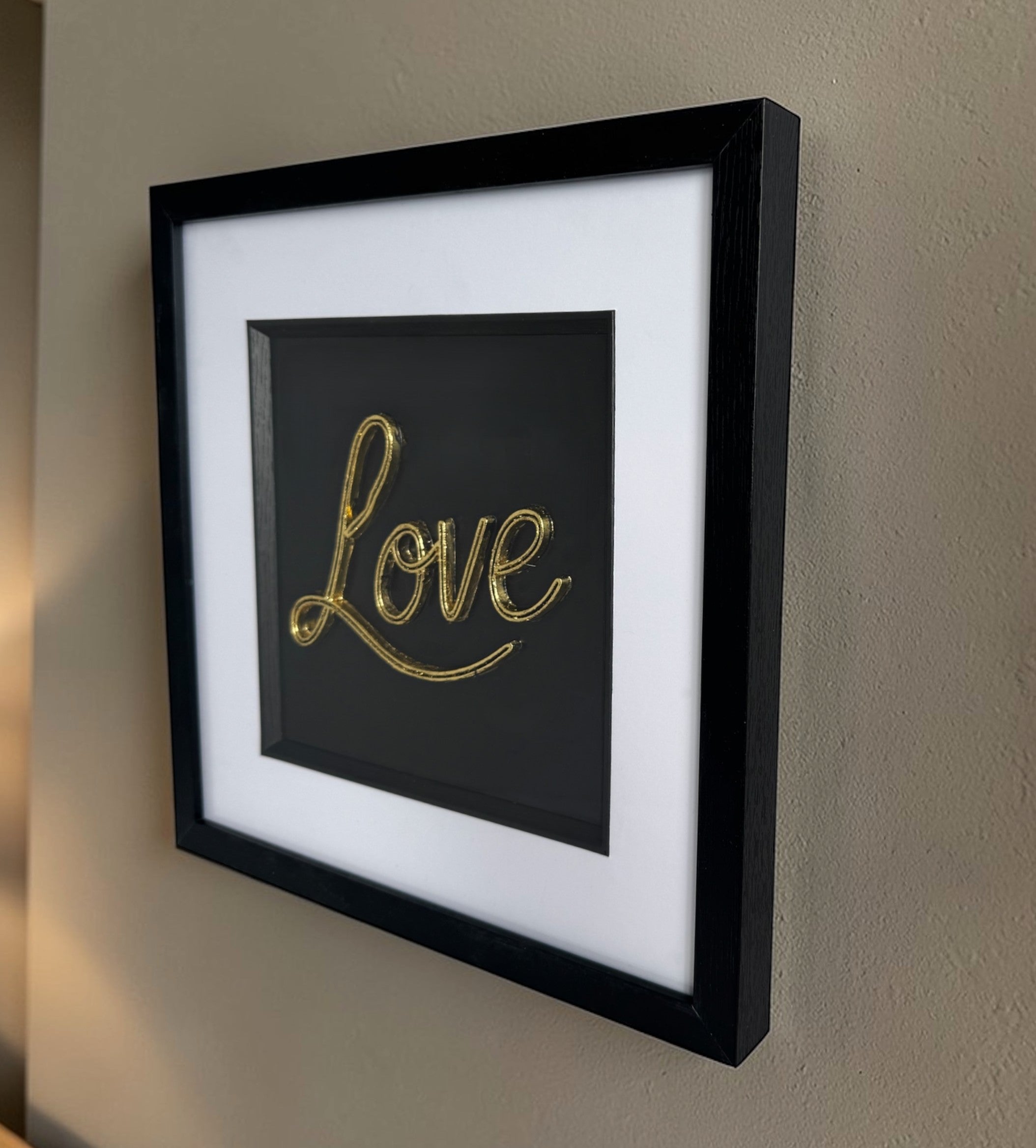 Angled wall display of framed 3D Love sign in gold finish with wide white mount and black inner surround.