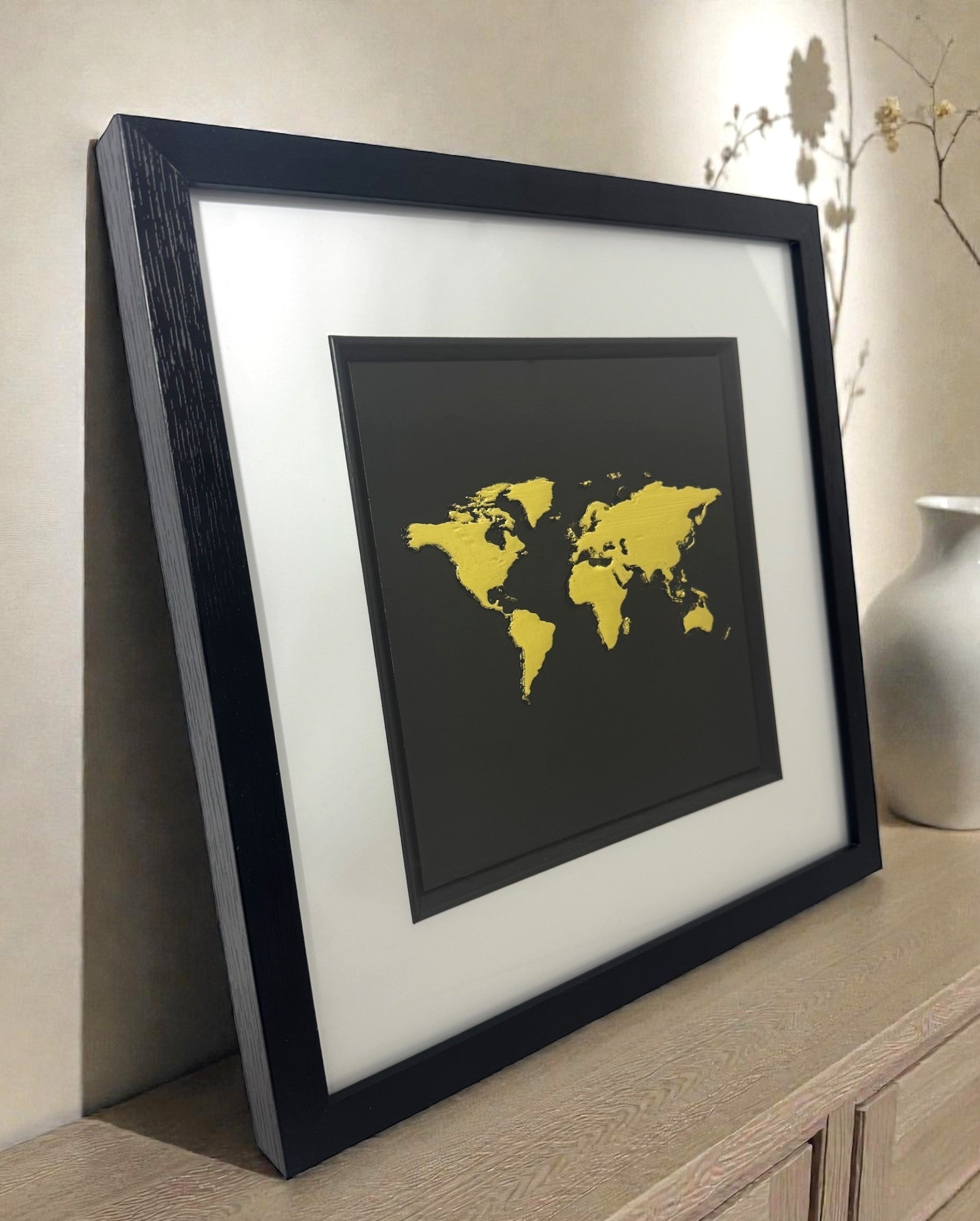 Large framed Gold World Map 3D art displayed on a shelf in a modern home. Luxury travel gift and metallic interior decor.