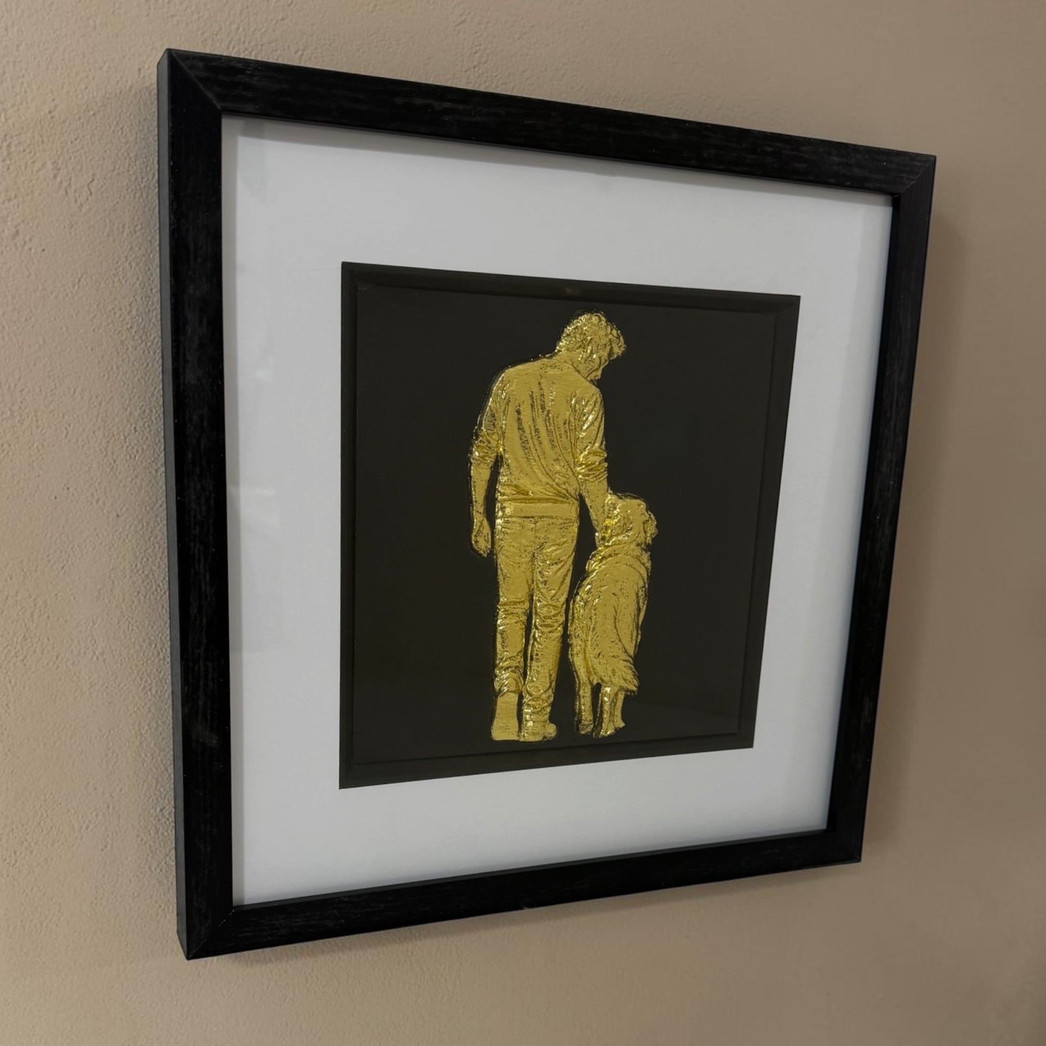 Angled view of gold man and dog relief artwork showing glass front, black box frame and white mount.