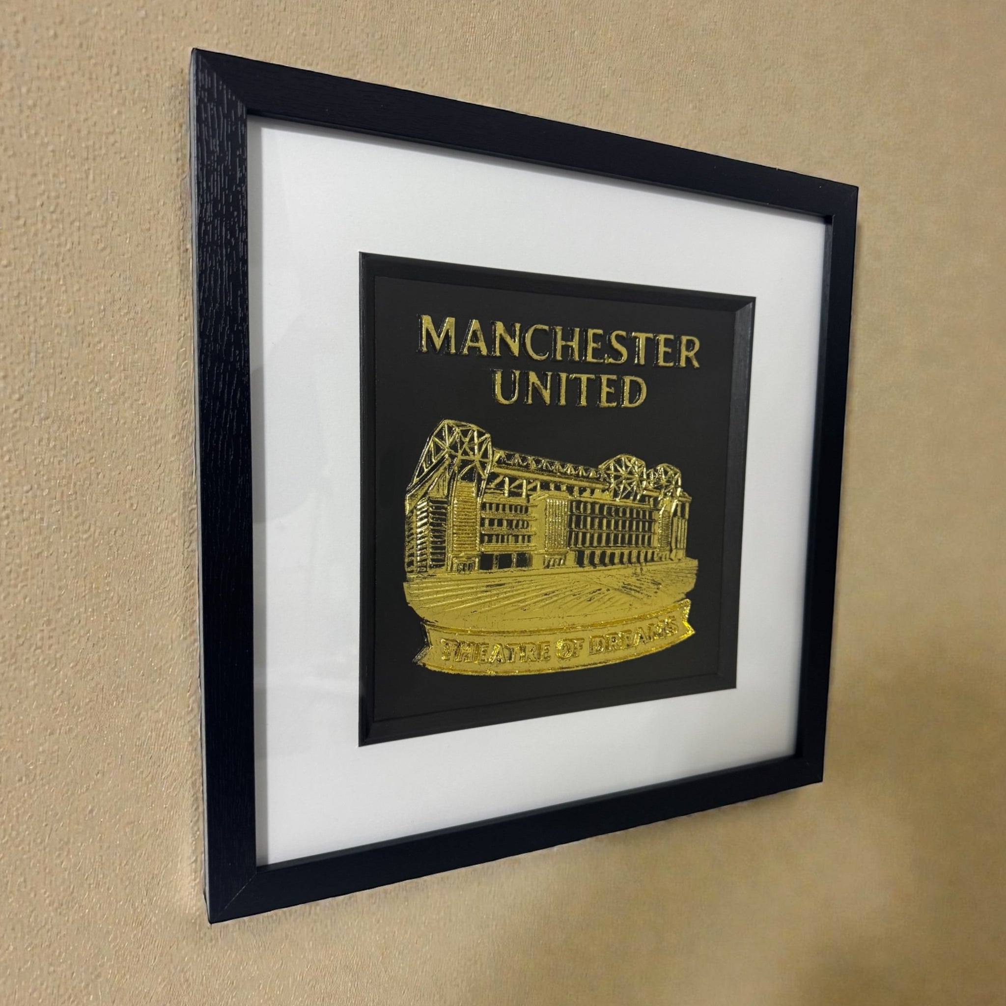 Manchester United gold relief framed wall art hanging on a wall, black frame and white mount, stadium artwork with “Theatre of Dreams” lettering.