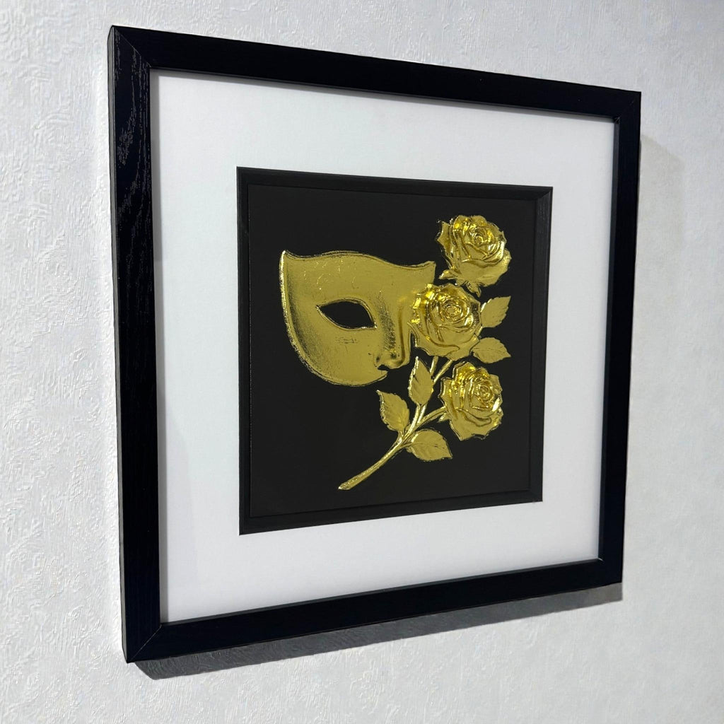 Masquerade mask and roses wall art in gold leaf hanging on a light textured wall in a black frame with white mount