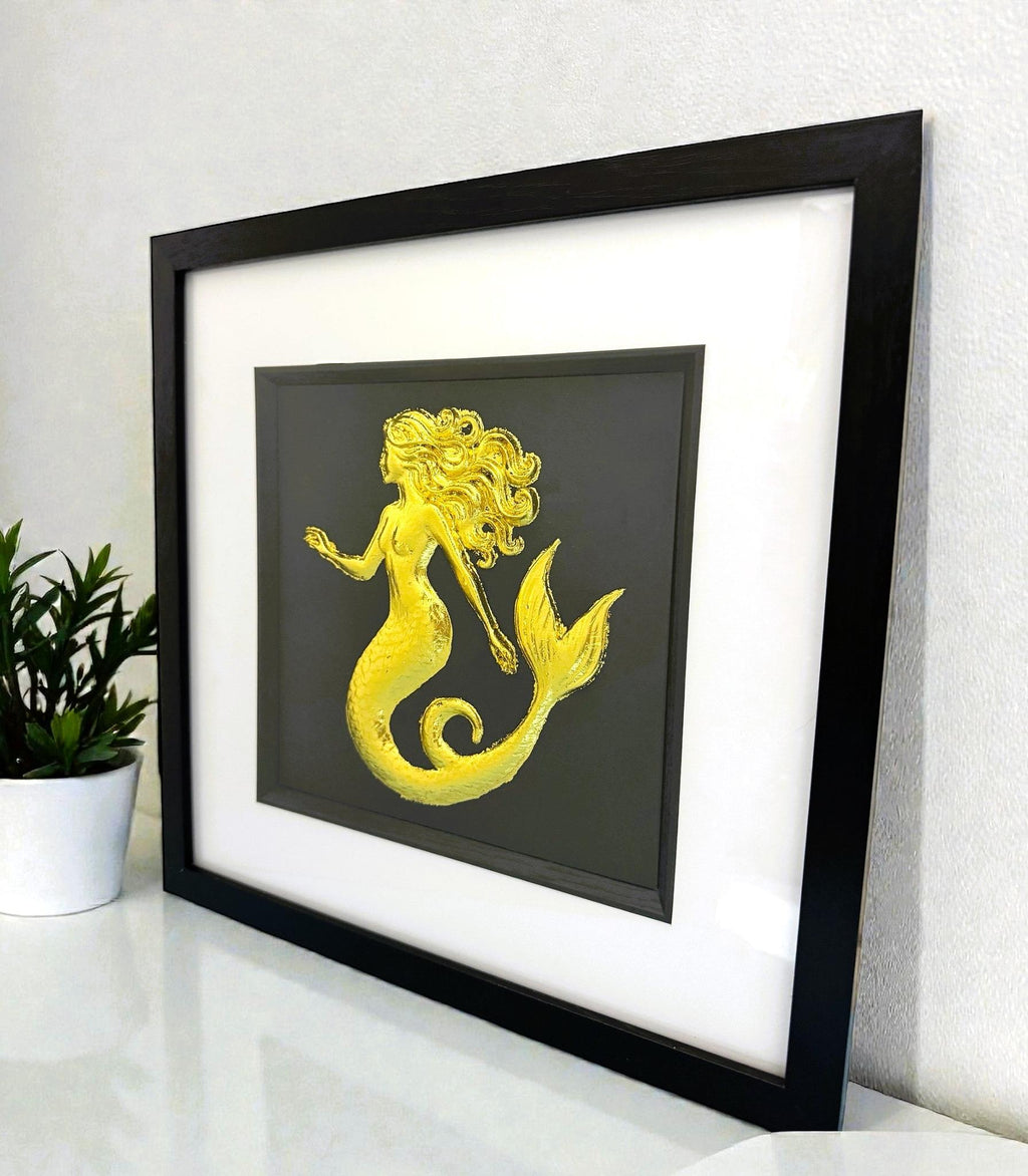 Gold mermaid wall art framed in black with white mount displayed on a shelf