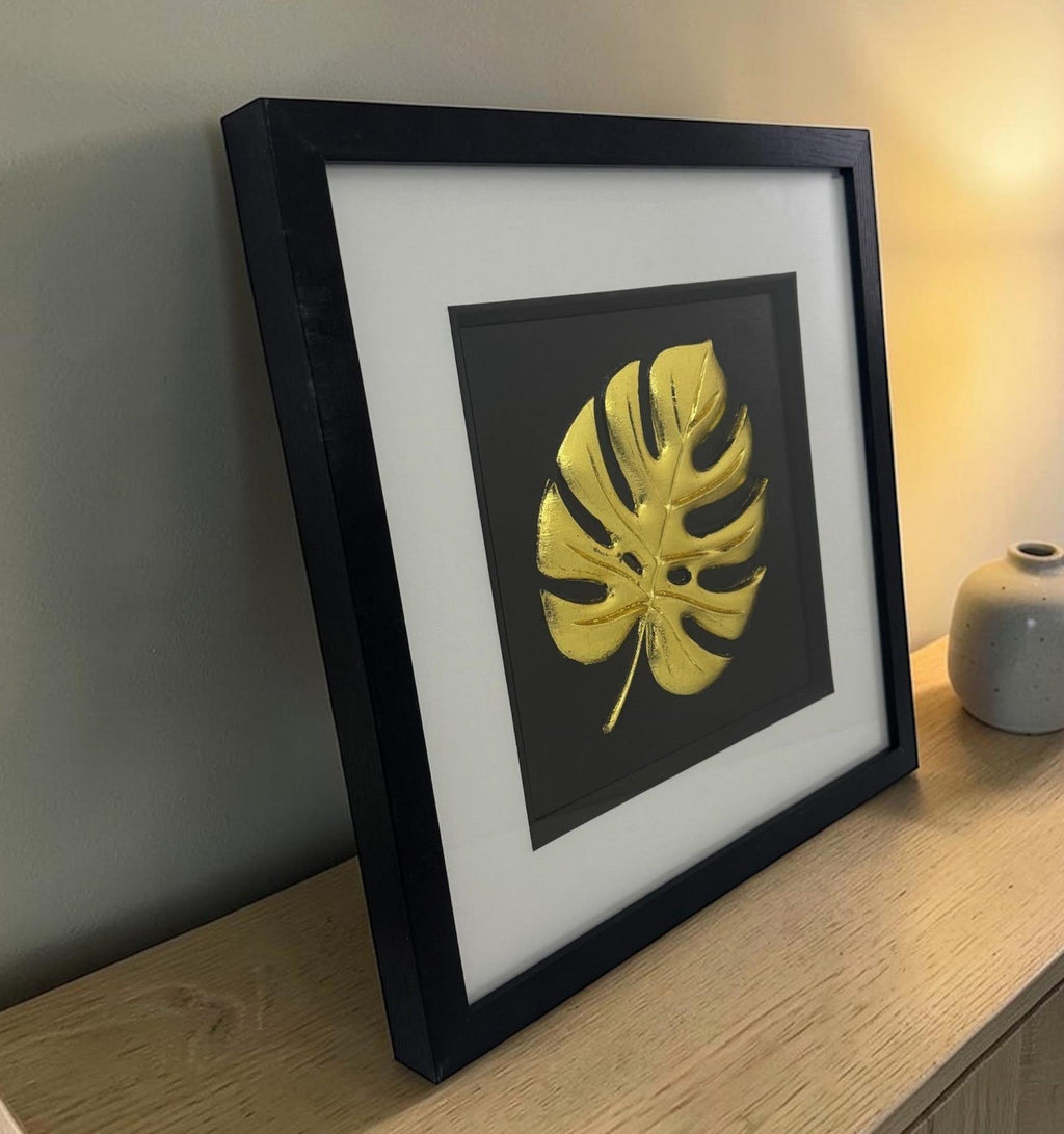 Gold monstera leaf 3D relief in black box frame with white mount, styled on a wooden sideboard