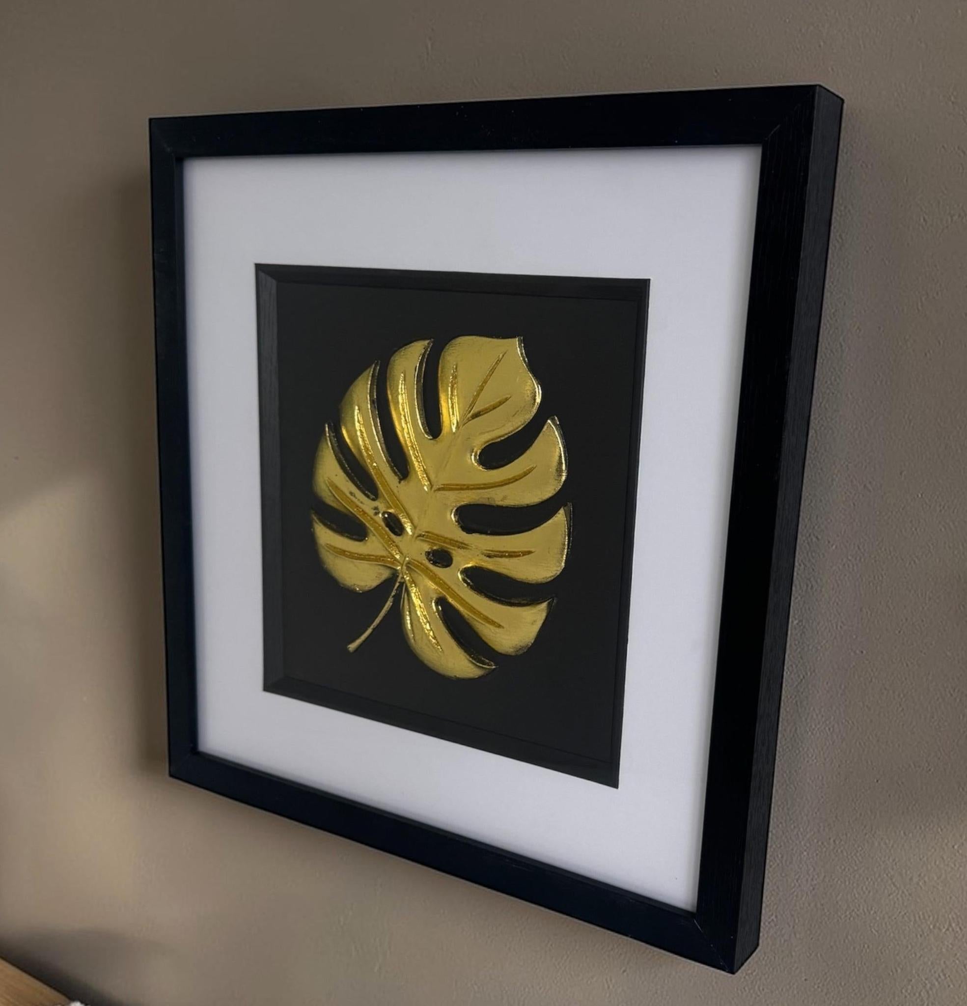 Gold monstera leaf relief artwork in square black box frame with wide white mount, hung on a neutral wall