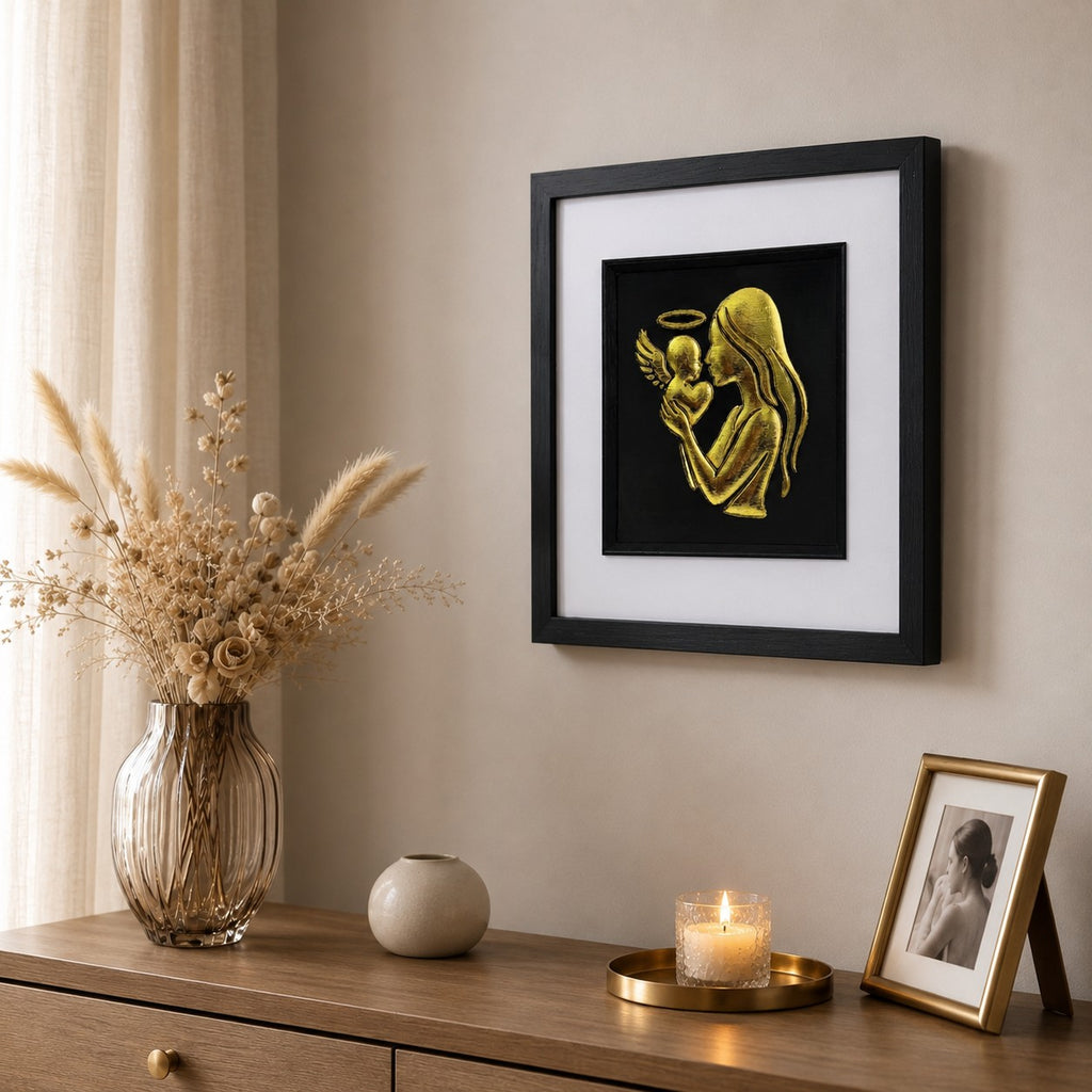 Angled lifestyle photo of framed mother and angel baby memorial wall art in gold leaf, shown in a calm neutral interior with dried flowers, candlelight and warm gold decor.