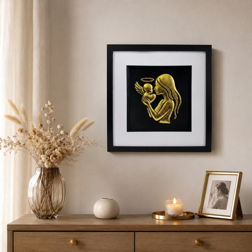 Framed mother and angel baby memorial wall art finished in gold leaf, displayed on a neutral wall above a wooden console with soft flowers, candlelight and gold accents.