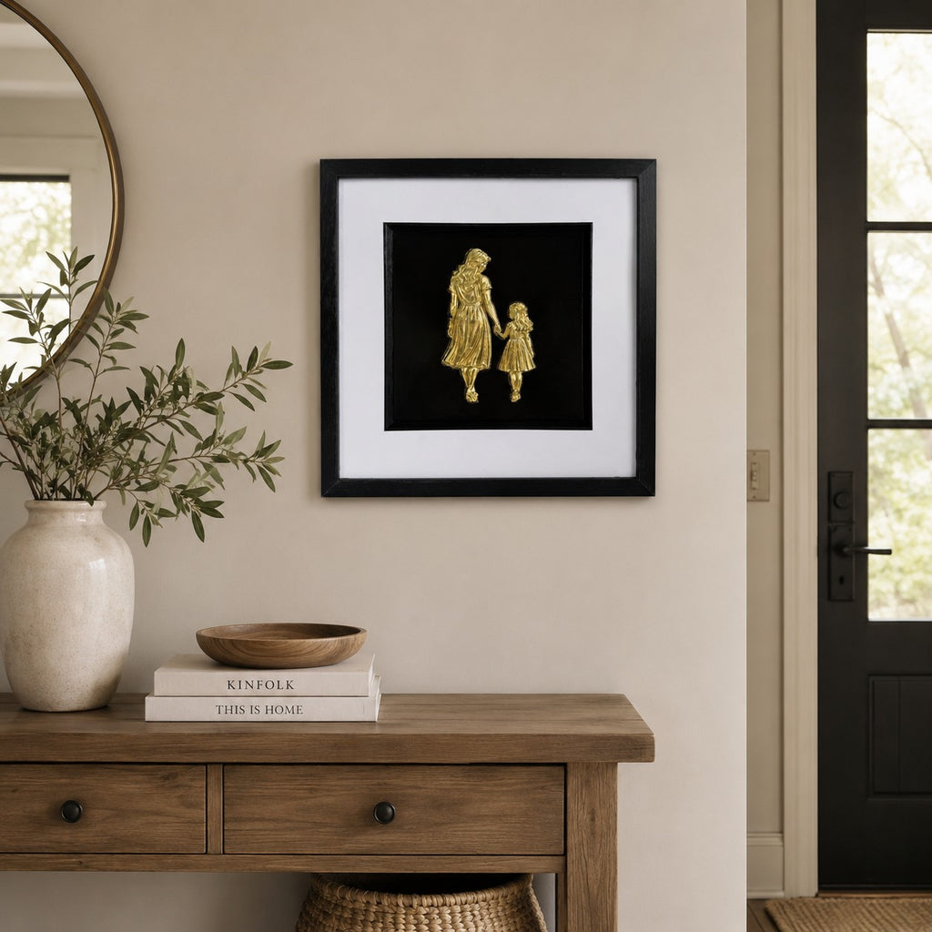 Gold mother and daughter framed artwork above wooden console with neutral decor and greenery.