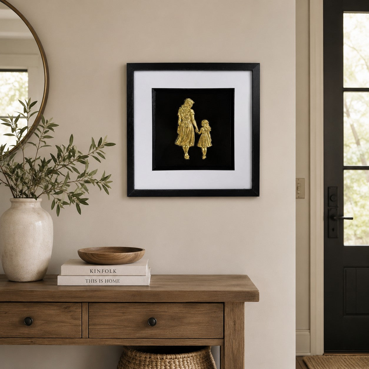 Gold mother and daughter framed artwork above wooden console with neutral decor and greenery.