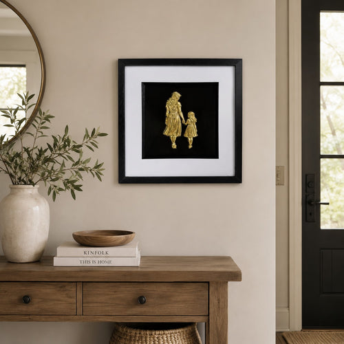 Gold mother and daughter framed artwork above wooden console with neutral decor and greenery.
