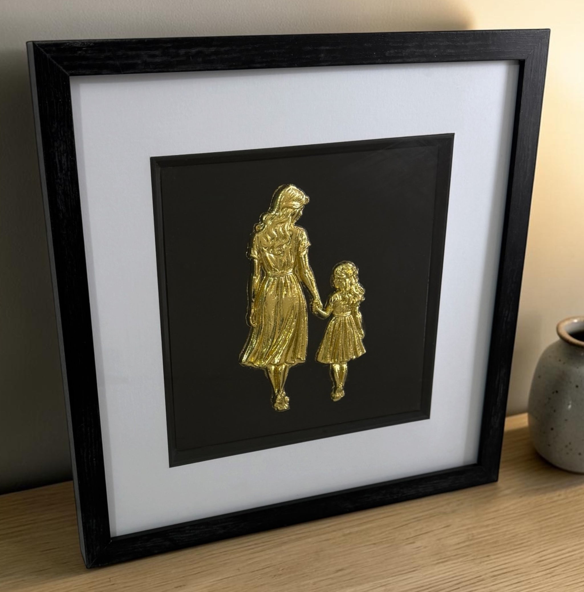 Gold mother and daughter 3D relief artwork in black frame with white mount on tabletop.