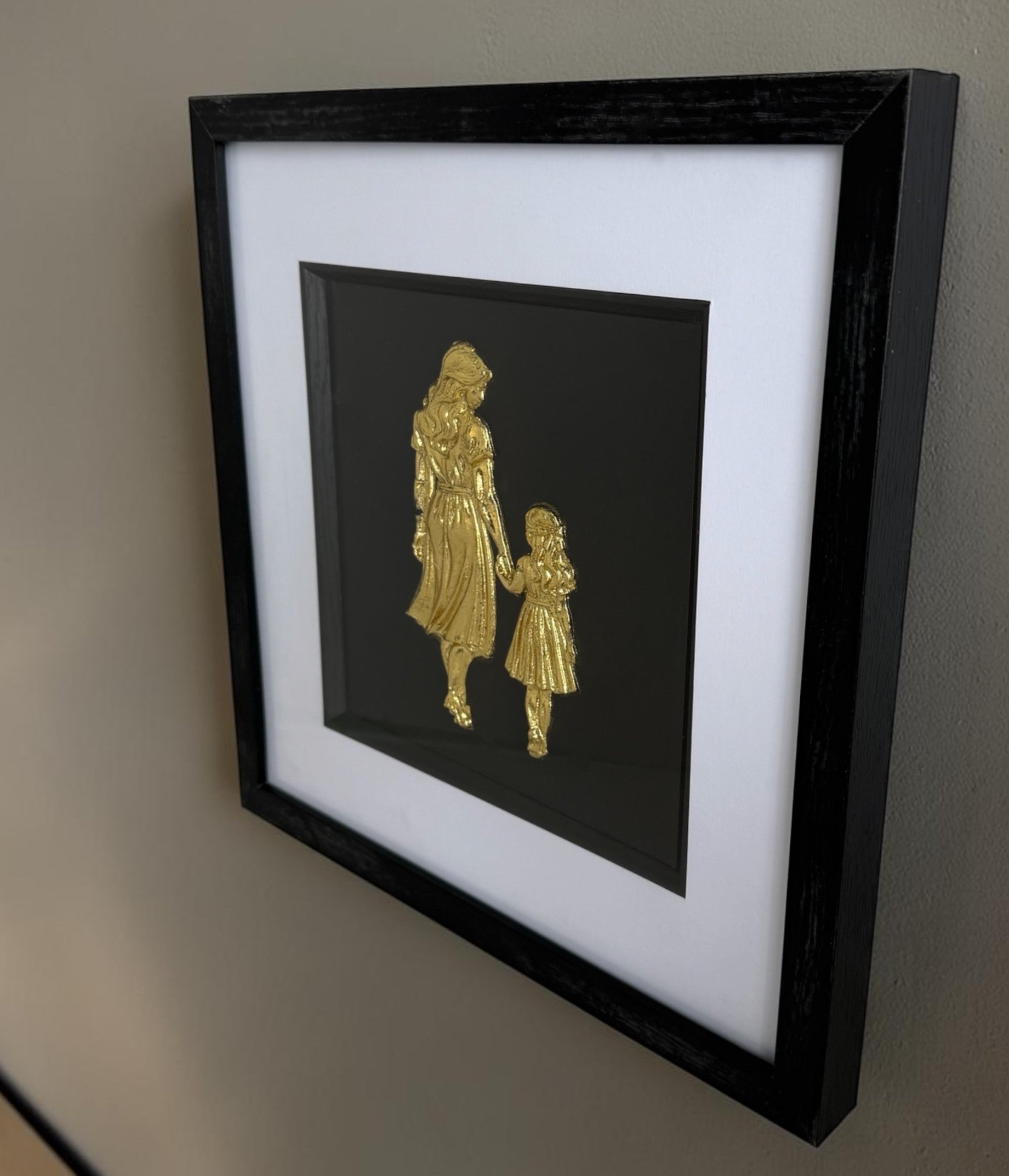 Angled wall view of Gold mother and daughter raised relief artwork showing black box frame depth.