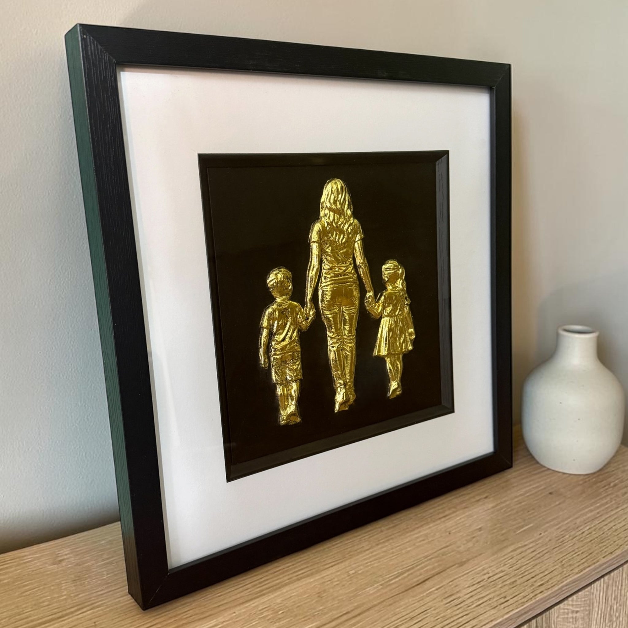 Mum, son and daughter framed wall art in gold leaf viewed at an angle on a sideboard, showing the raised relief depth in a black wooden box frame with white mount