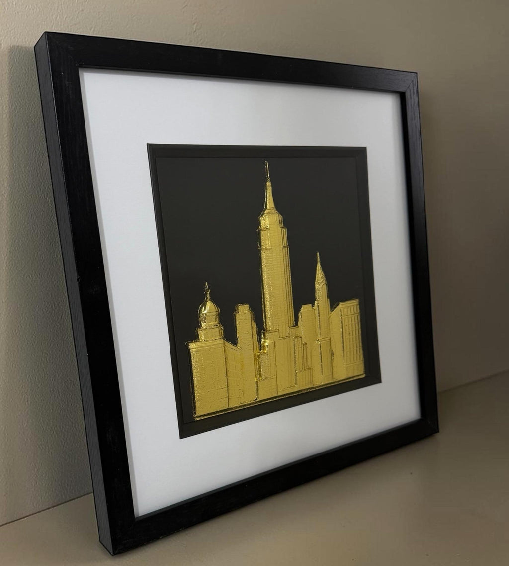 Gold framed New York skyline artwork leaning on a neutral surface with black box frame, white mount and glass front.
