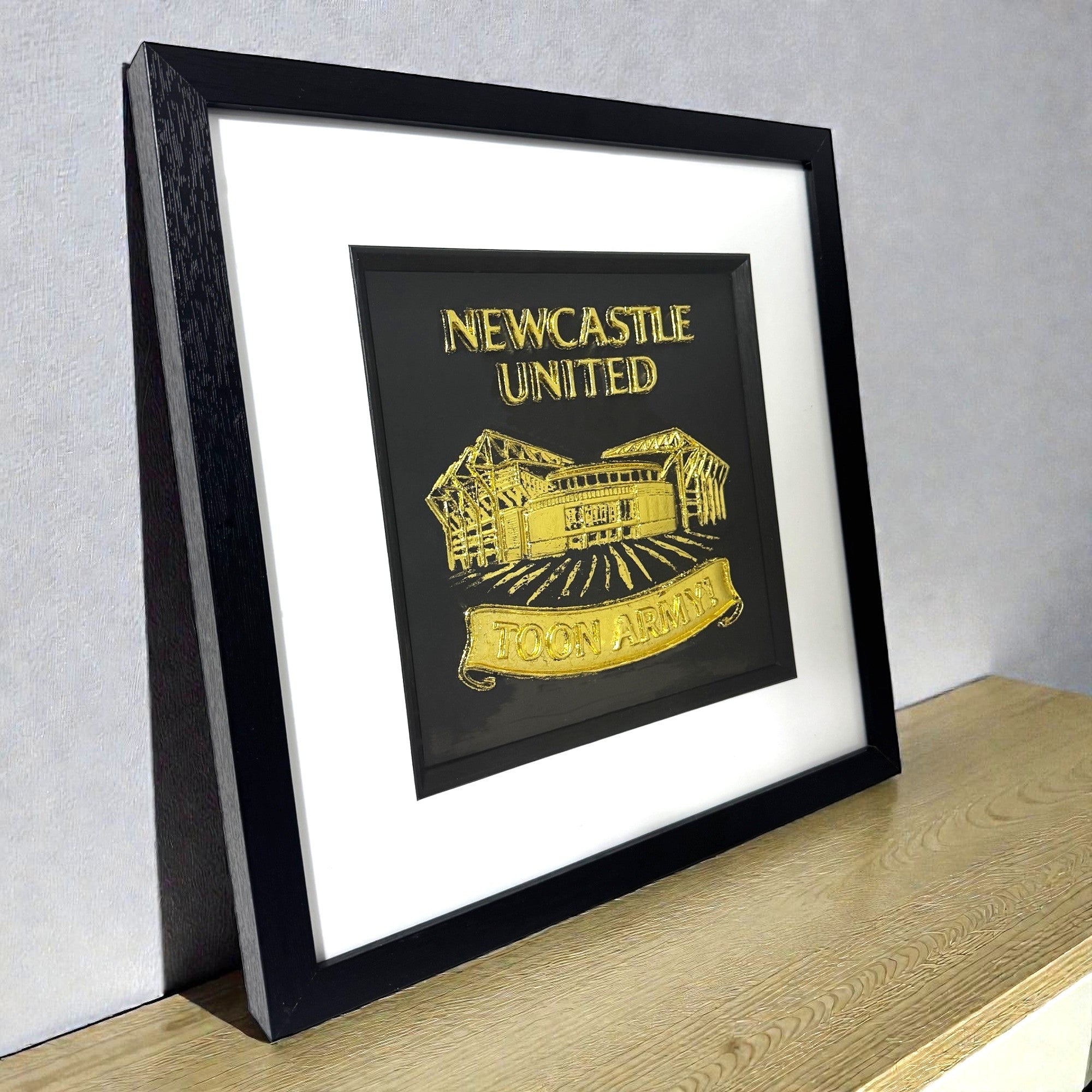 Framed Newcastle United gold 3D wall art displayed on a modern wooden shelf.