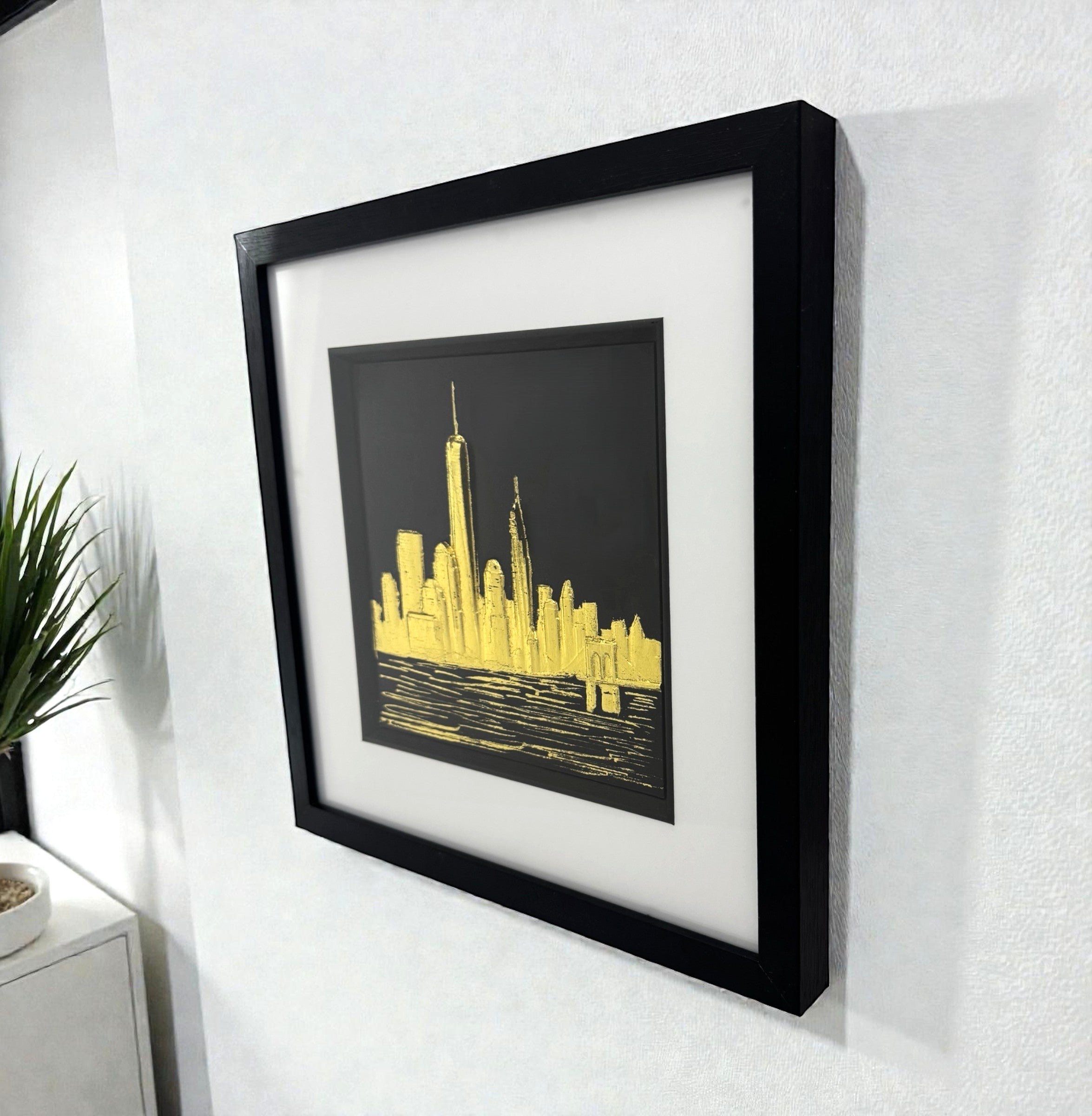 Gold leaf New York skyline wall art displayed on a wall, angled perspective showing scale, shadow depth, and framed finish