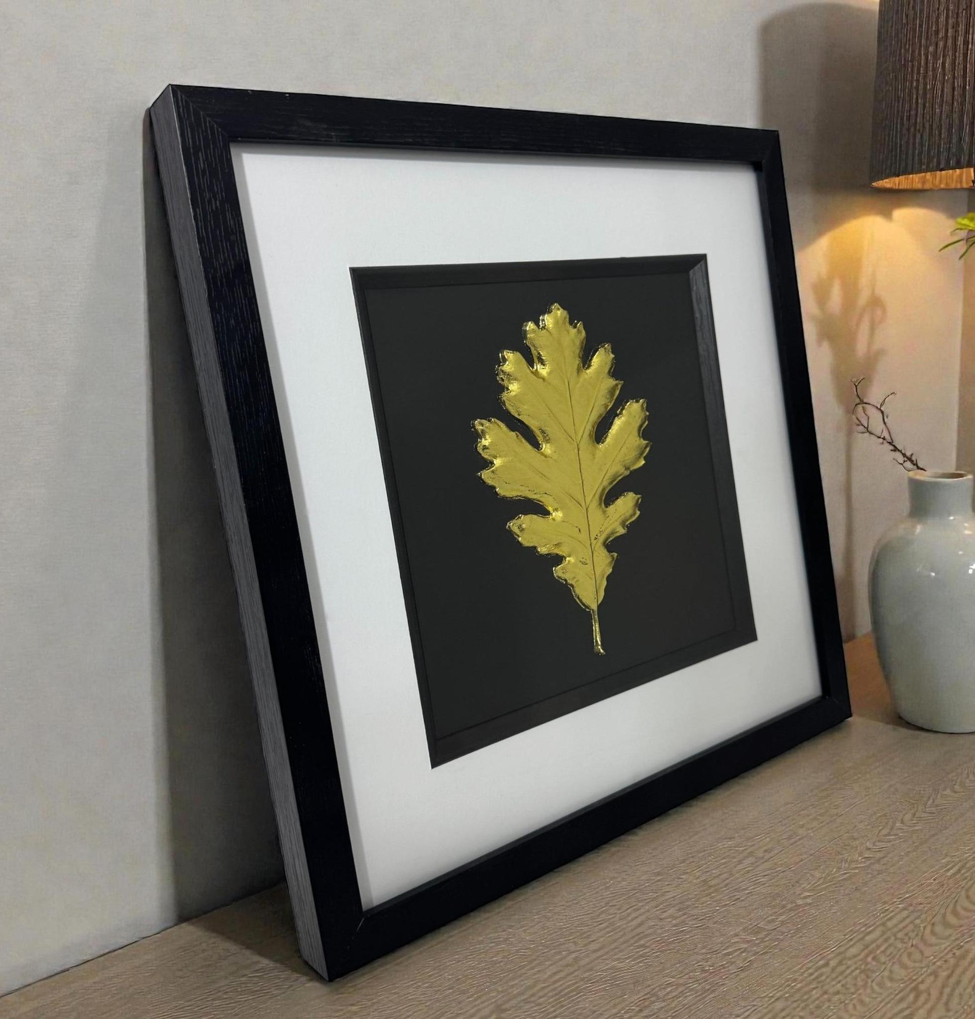 Framed oak leaf 3D relief artwork in Gold Leaf, black box frame with white mount and inner black surround, styled on a wooden sideboard