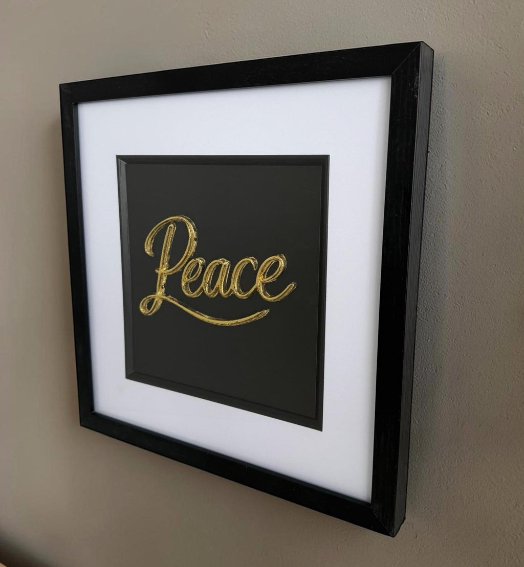 Angled gold Peace word artwork on a wall showing raised lettering, black box frame depth and glass front.