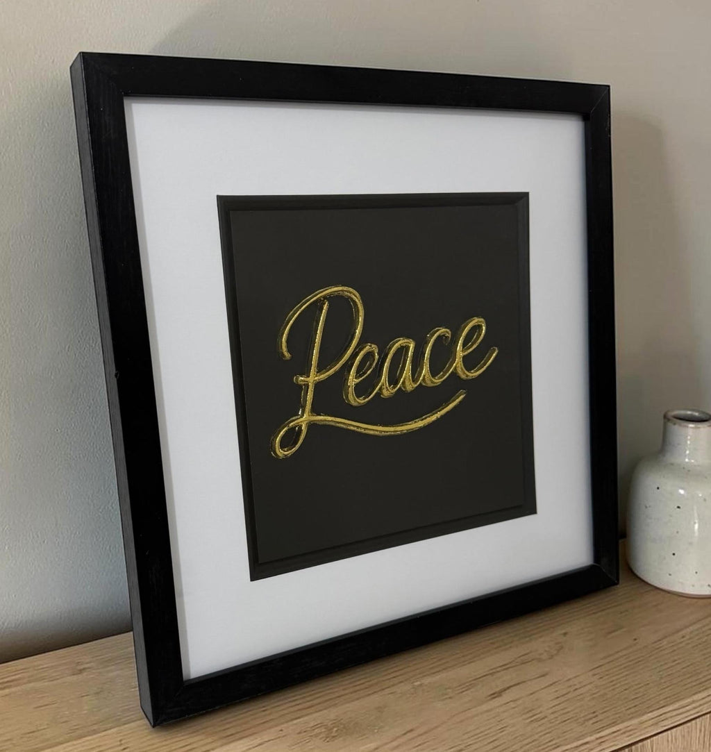 Gold framed Peace word artwork leaning on a wooden sideboard beside a small ceramic vase.