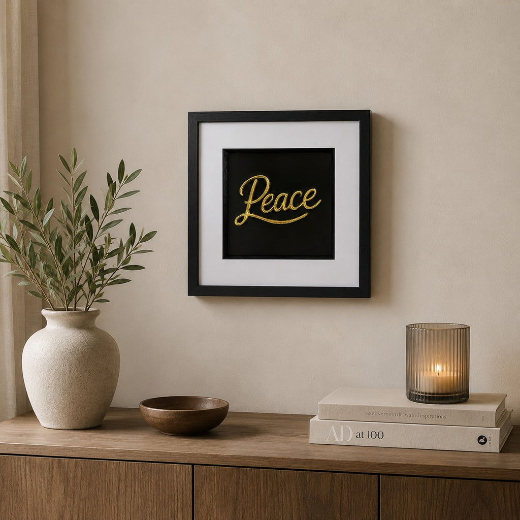 Gold framed Peace word artwork styled on a neutral wall above a wooden sideboard with vase, candle and books.