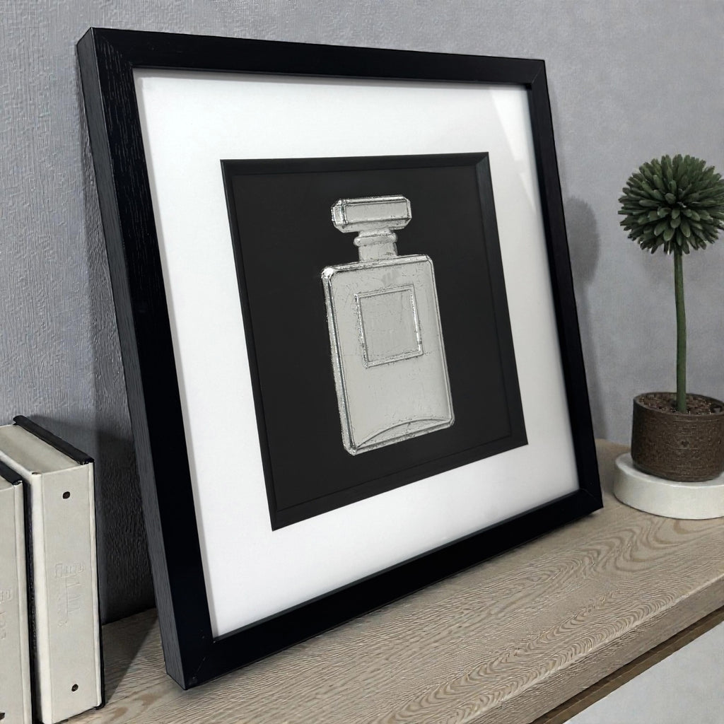 Framed perfume bottle wall art in silver leaf displayed on a shelf, black frame with white mount and raised relief design