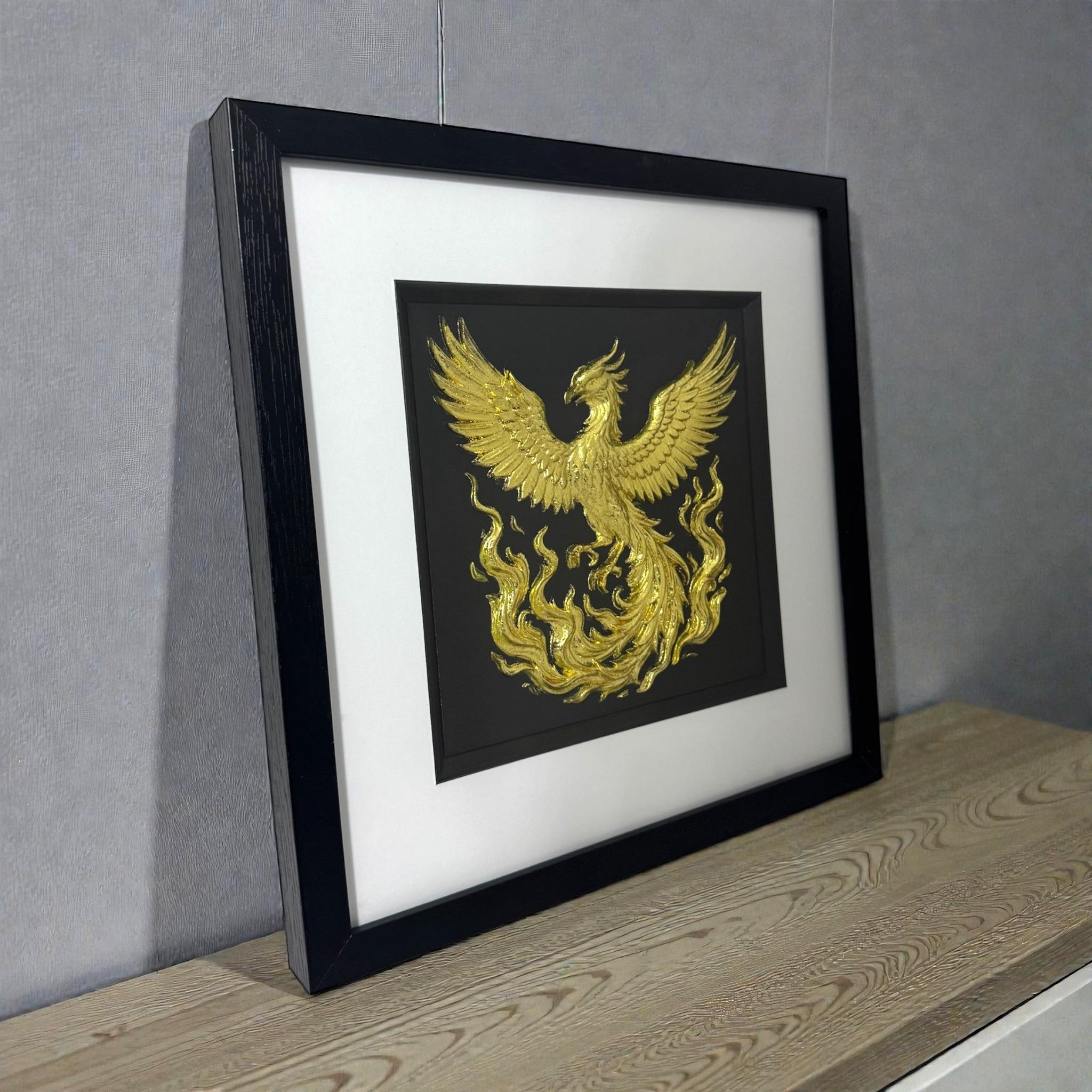 Gold phoenix rising wall art shown at an angle on a wooden surface, framed with white mount and raised phoenix relief catching the light