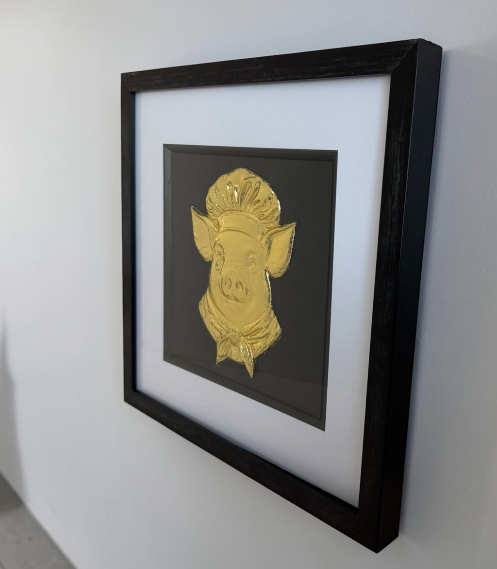 Angled wall view of gold pig chef raised artwork in black box frame with white mount and glass front