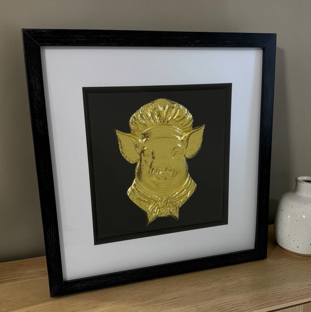 Gold pig chef framed artwork leaning on a wooden surface with neutral ceramic vase styling