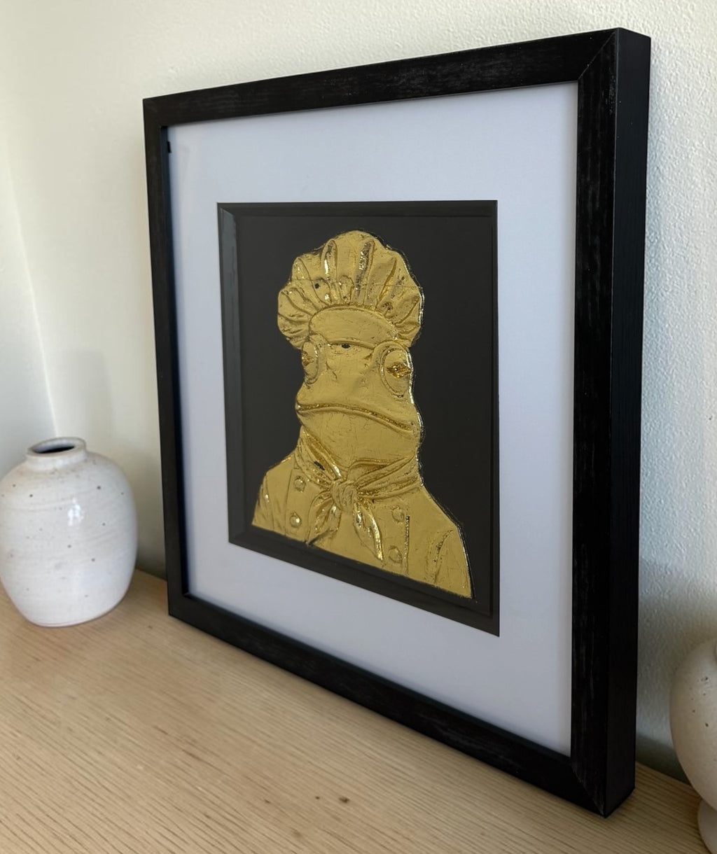 Gold frog chef raised artwork in black box frame on a wooden surface with white ceramic vases