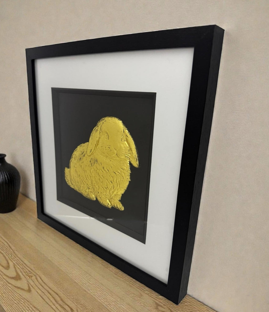 Side view of framed Mini Lop rabbit 3D wall art in gold leaf, in a black frame with white mount, styled on a wooden surface.