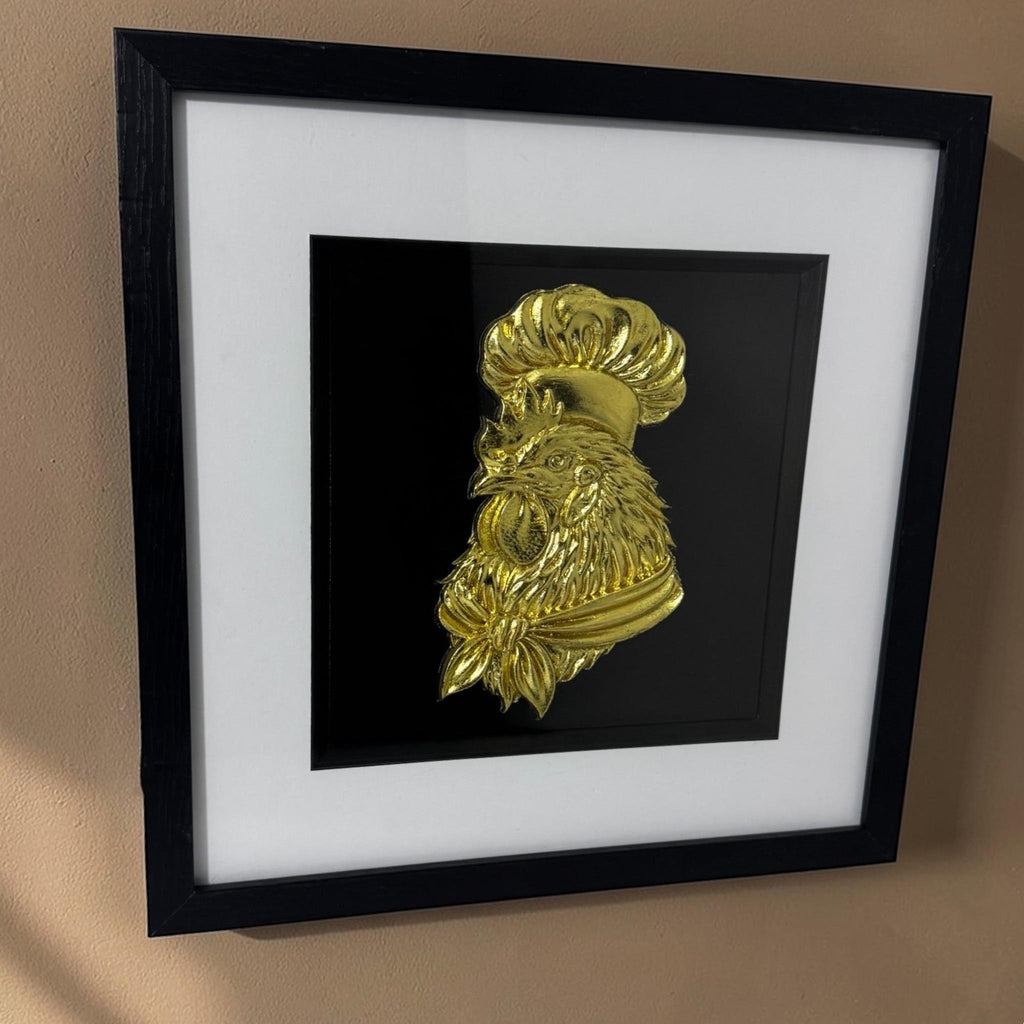 Angled view of a gold chef rooster relief in a black box frame hanging on a beige wall.