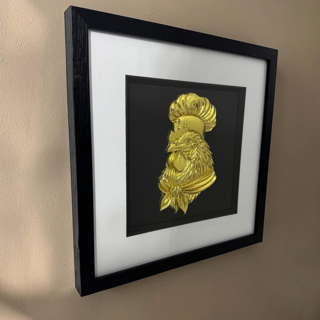 Angled view of a gold chef rooster relief in a black box frame hanging on a beige wall with warm side lighting.