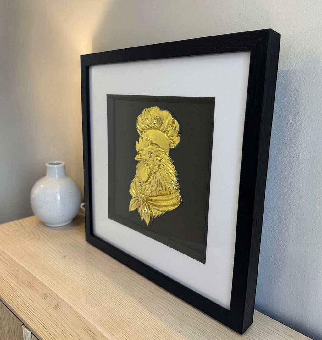 Gold chef rooster relief in a black box frame with wide white mount, styled on a wooden sideboard beside a white ceramic vase.