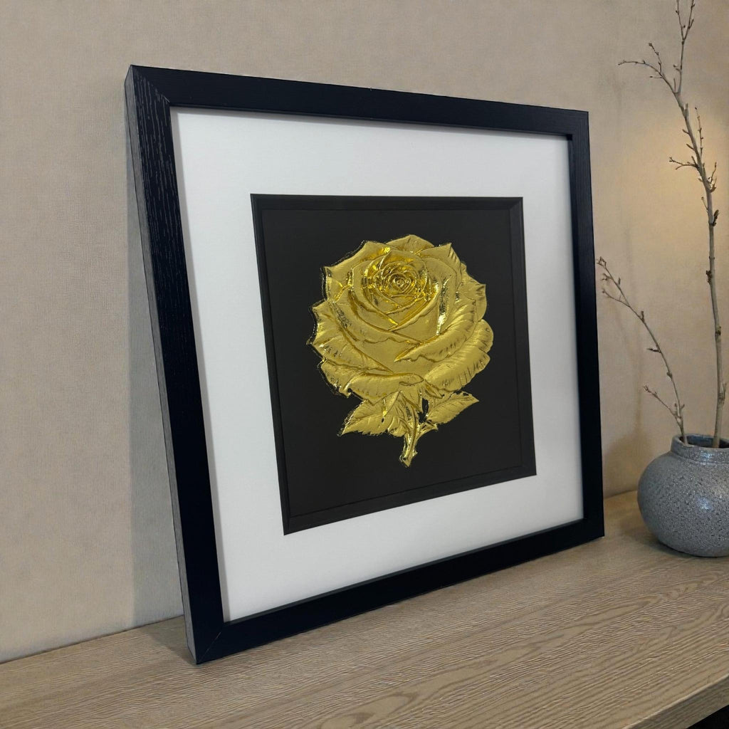 Angled lifestyle photo of framed 3D rose wall art in Gold Leaf, in a deep black box frame with crisp white mount on a wooden surface.