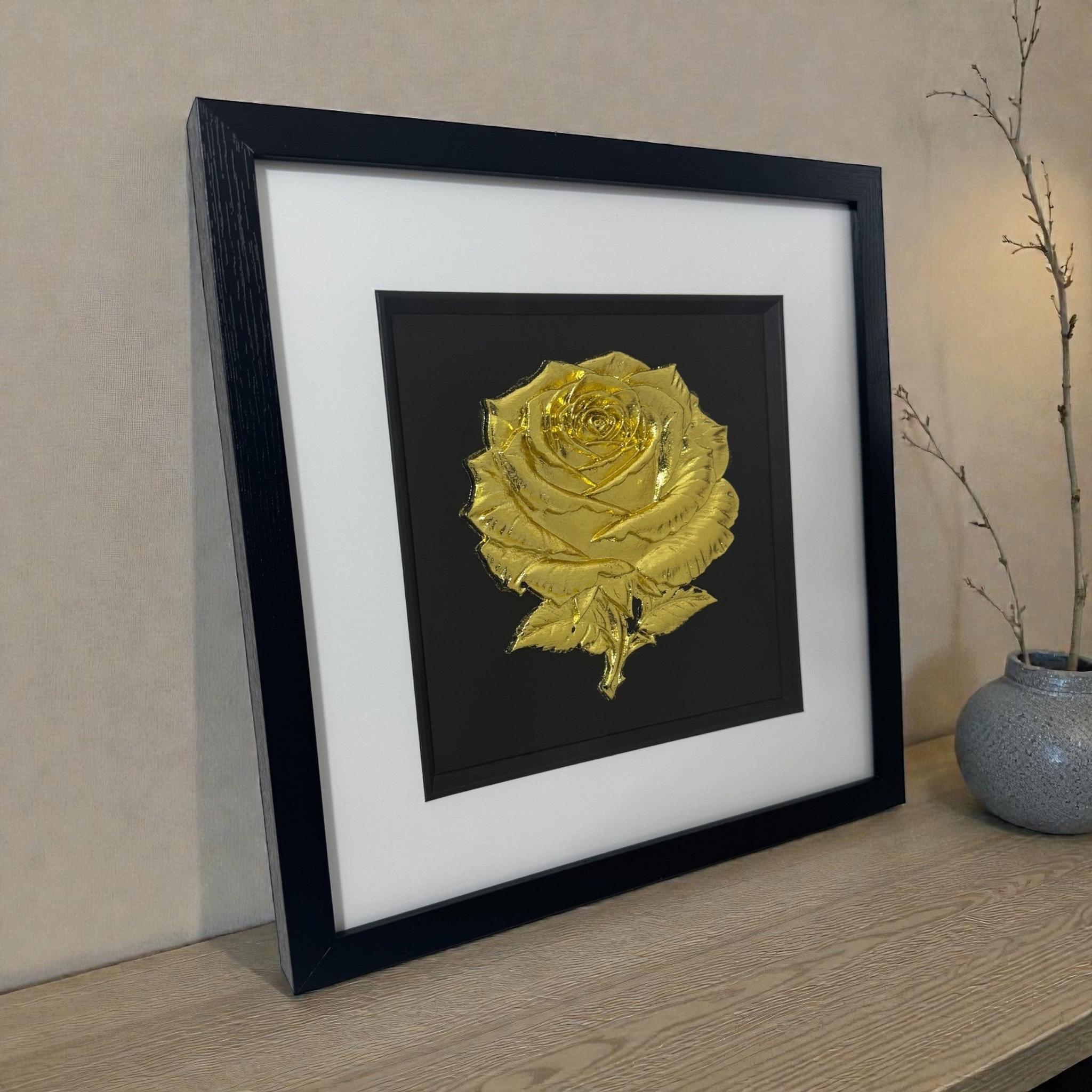 Angled lifestyle photo of framed 3D rose wall art in Gold Leaf, in a deep black box frame with crisp white mount on a wooden surface.