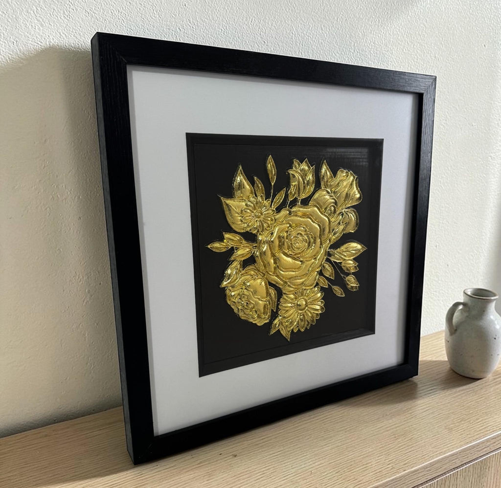 Angled view of framed gold-finish floral bouquet relief on a light wood sideboard beside a small ceramic vase.