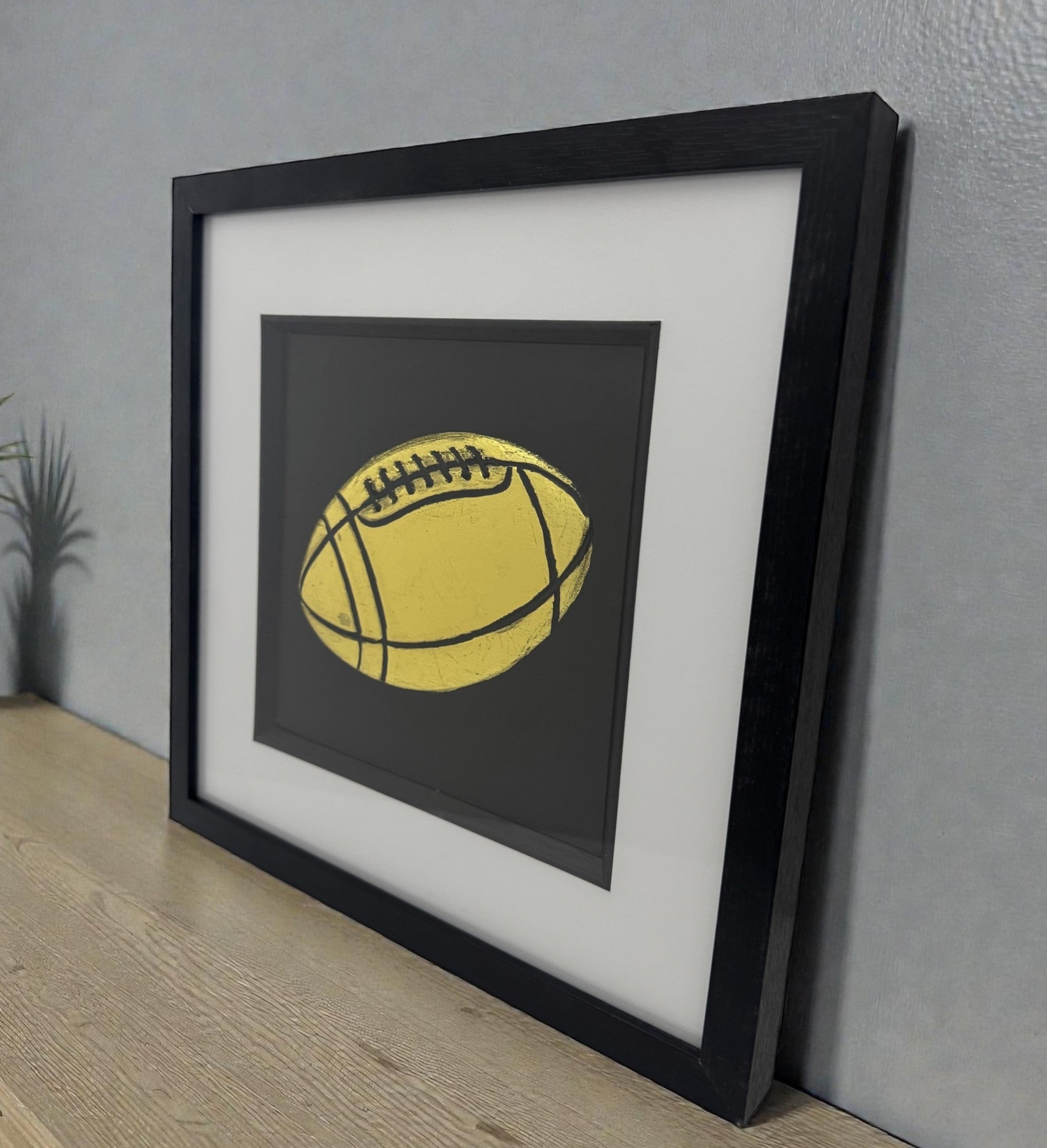 Rugby ball relief artwork finished in Gold Leaf on black background, framed with white mount, styled on a wooden sideboard