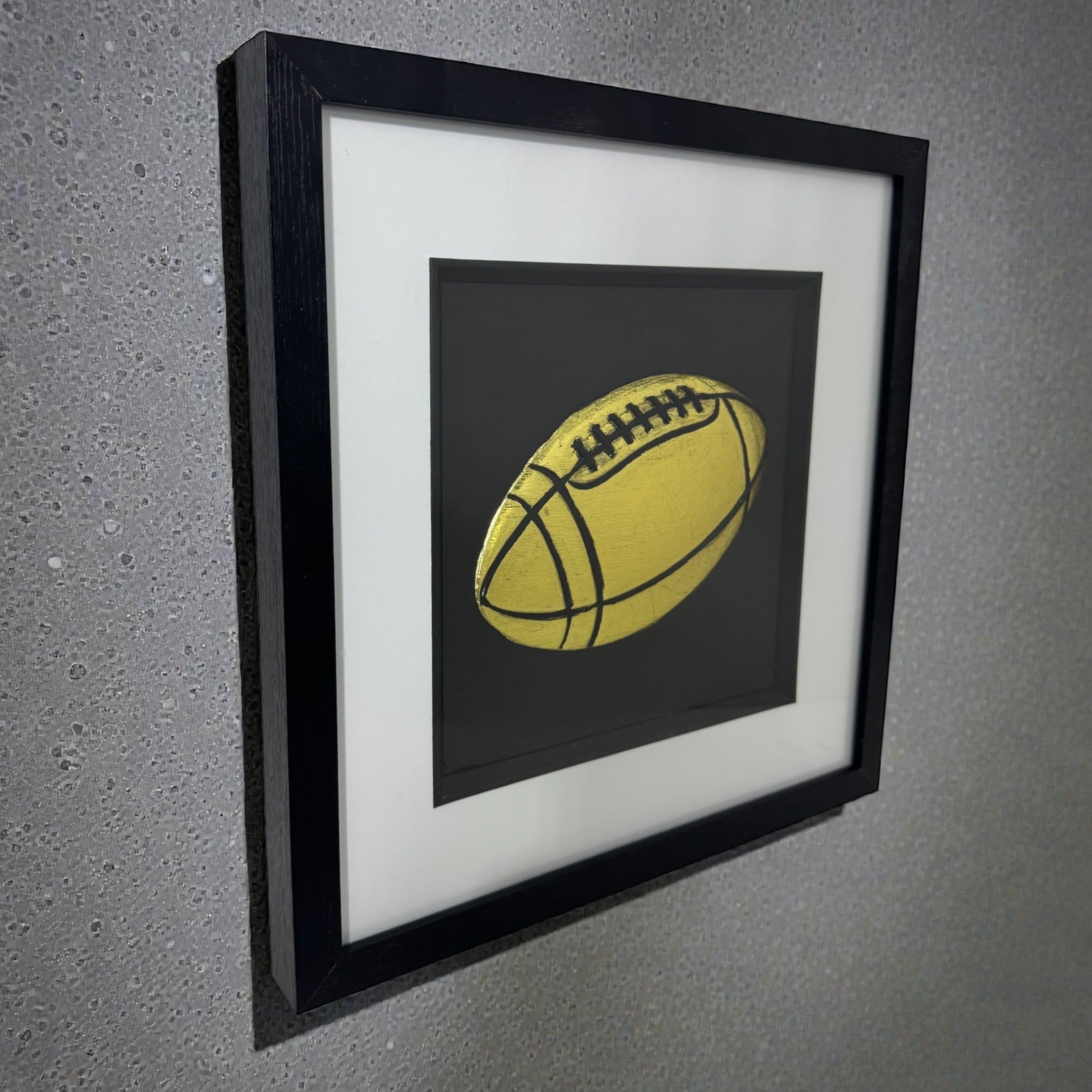 Framed rugby ball 3D relief artwork in Gold Leaf, black box frame with white mount, angled on a grey wall
