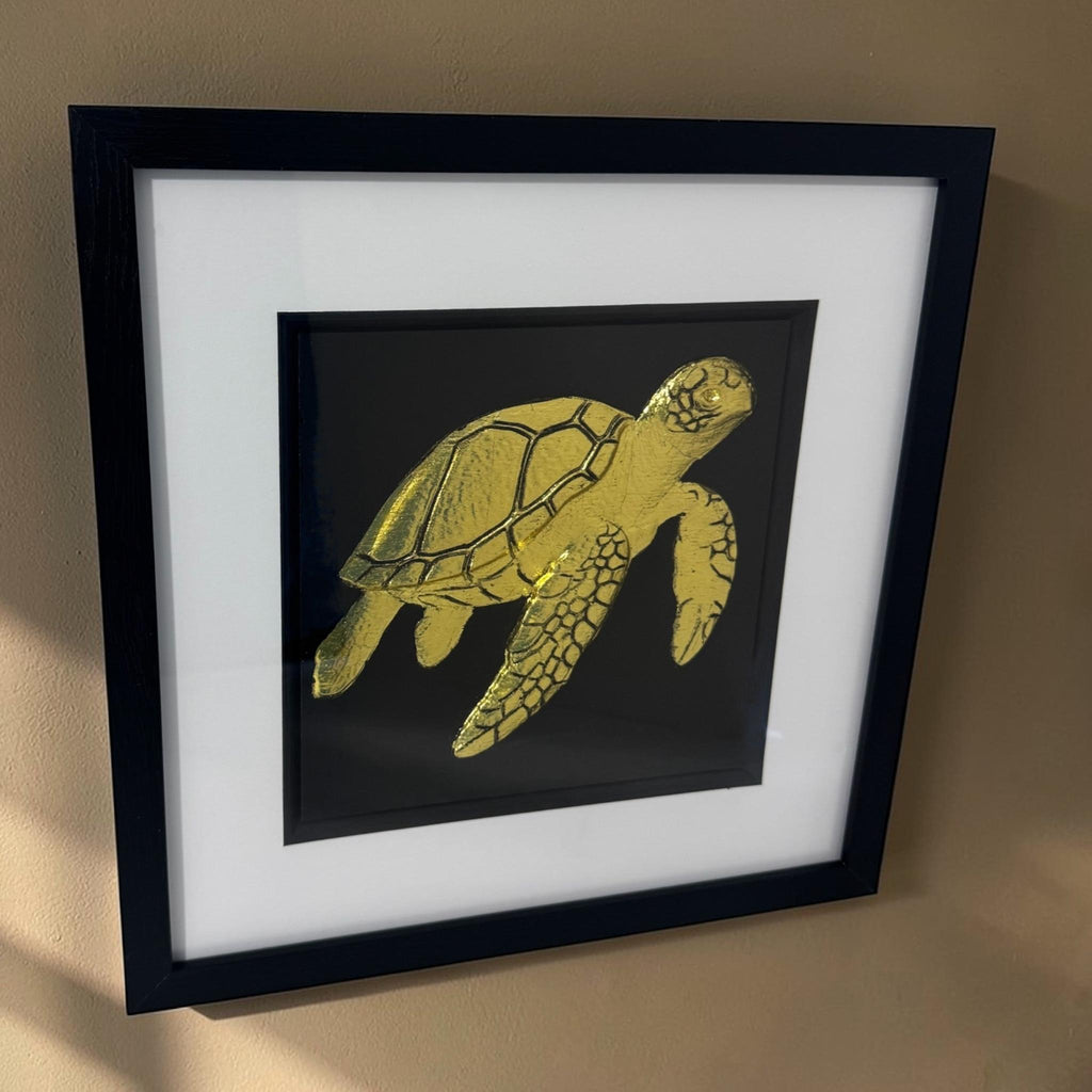 Angled view of the gold framed sea turtle relief with glass front on a neutral wall in warm light