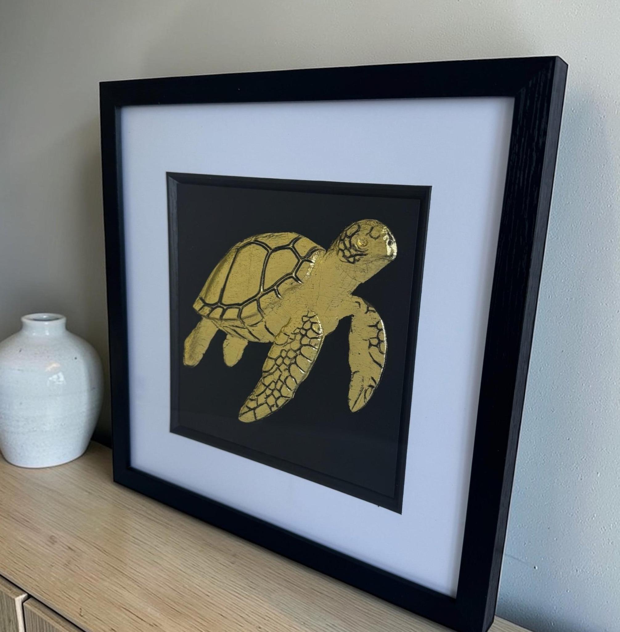 Gold sea turtle wall decor in a black frame with a white mount on a light wood sideboard beside a white vase