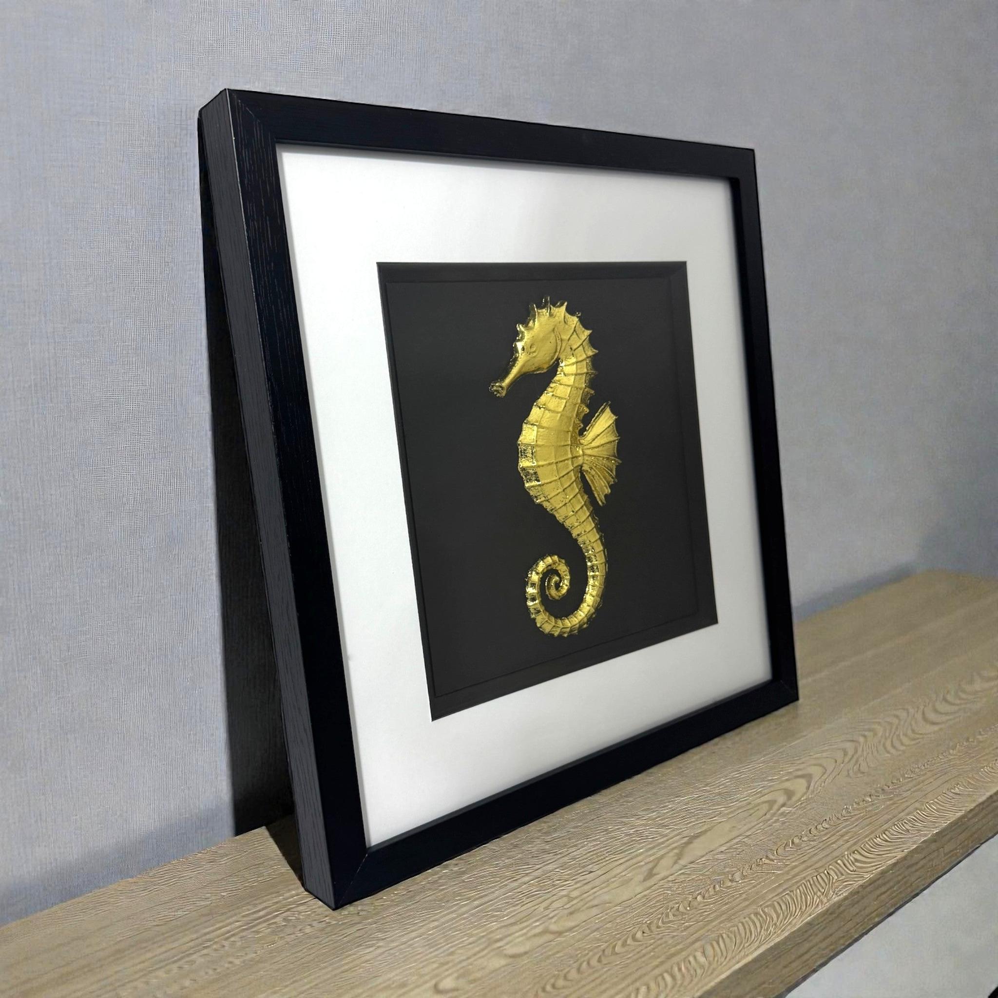 Gold seahorse wall art shown at an angle on a wooden surface, framed with white mount and raised seahorse relief