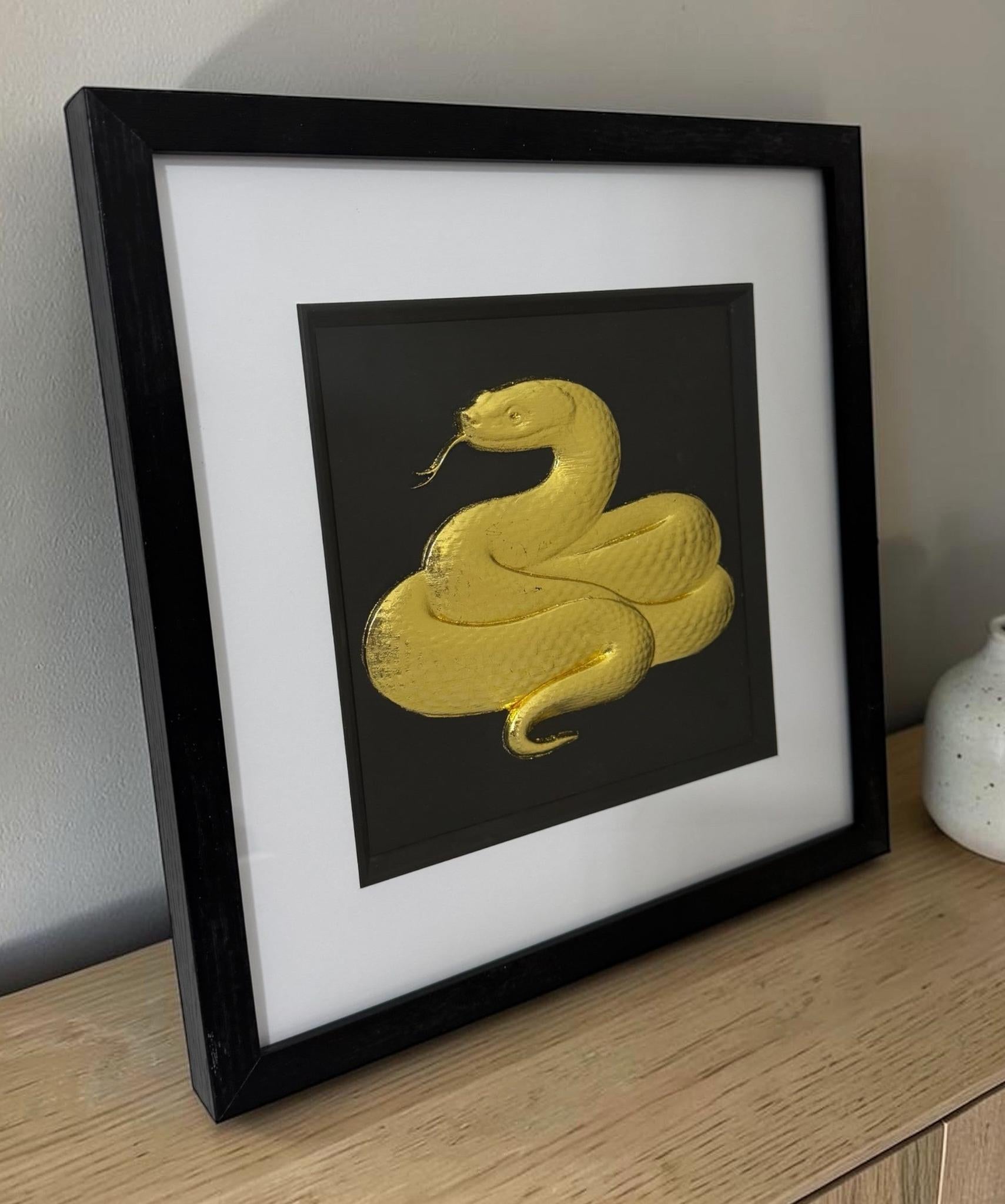 Gold framed snake wall art shown on a wooden sideboard with black frame, white mount and raised serpent relief.