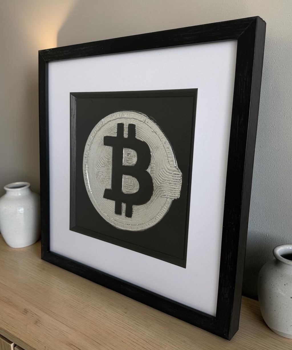 side view of Bitcoin silver leaf wall art in black frame on a sideboard against neutral wall