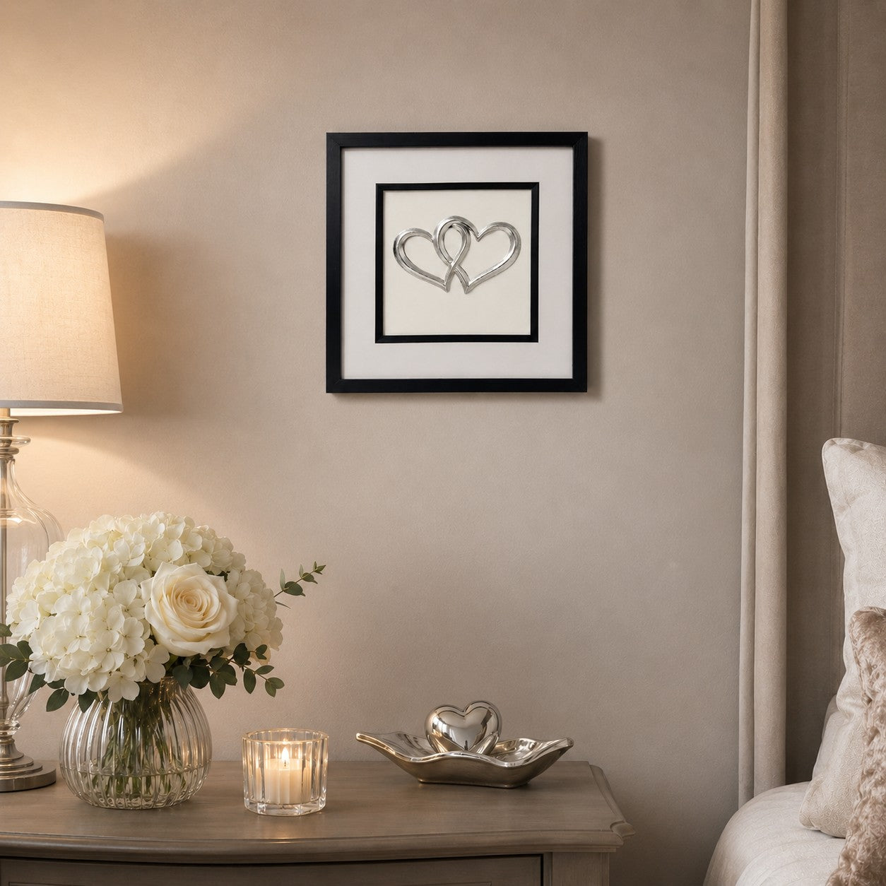 Silver joined hearts wall art in a black square frame styled above a neutral bedside table with white flowers, candlelight and soft bedroom decor.